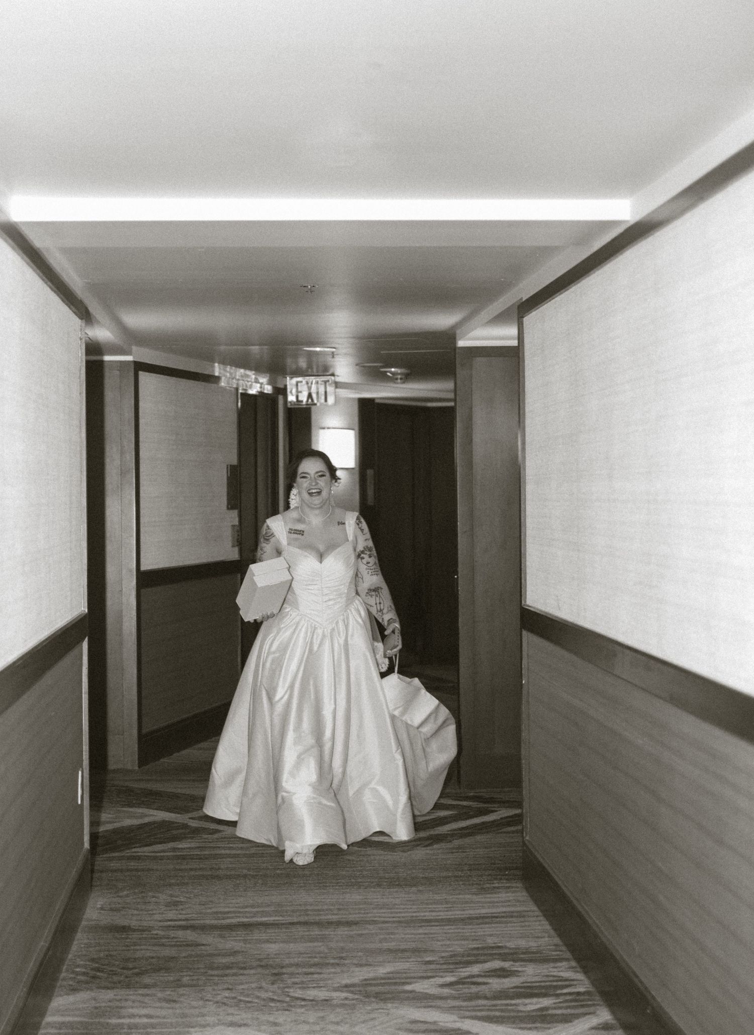 Bride in white wedding gown walking down hotel hallway, black and white photo.