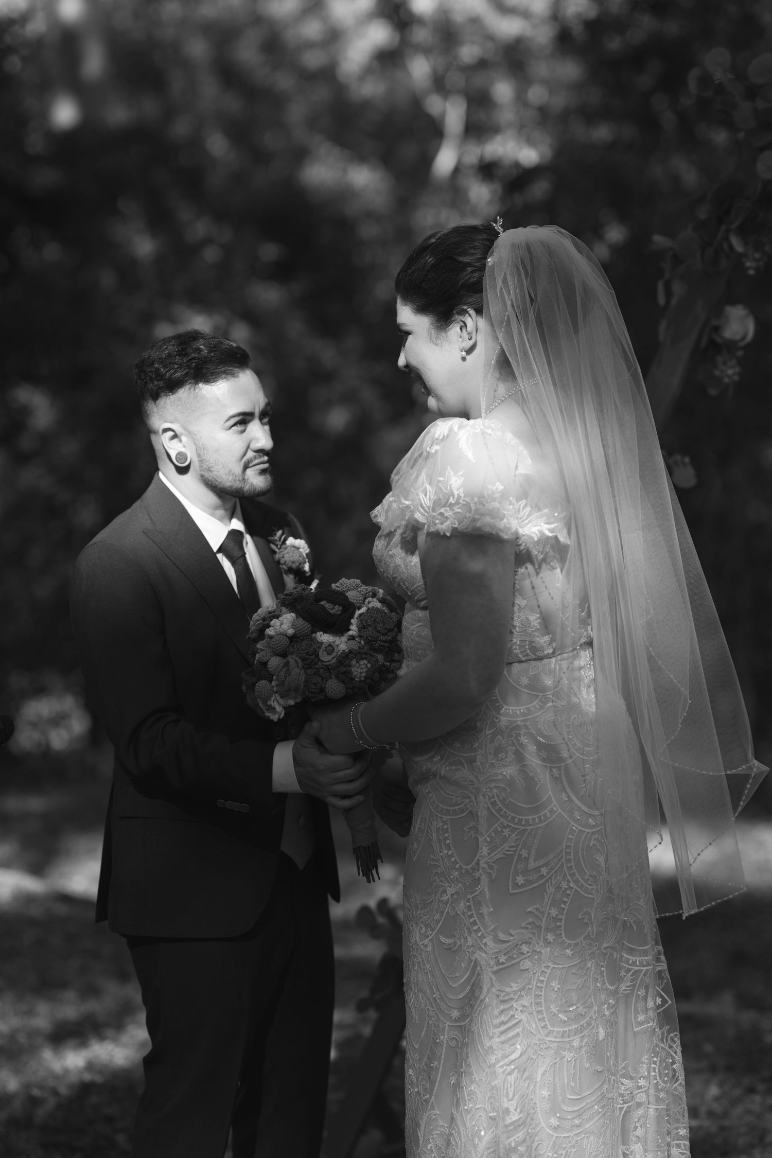 Black and white photo of a smiling bride in lace gown and veil holding bouquet with groom in suit outdoors.
