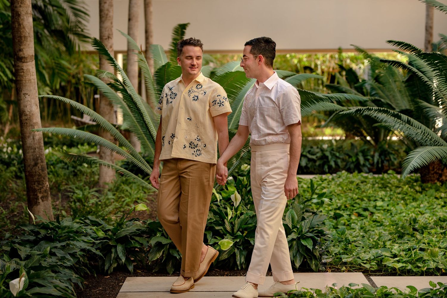 Two men holding hands and walking together through a lush tropical garden, wearing stylish casual outfits.