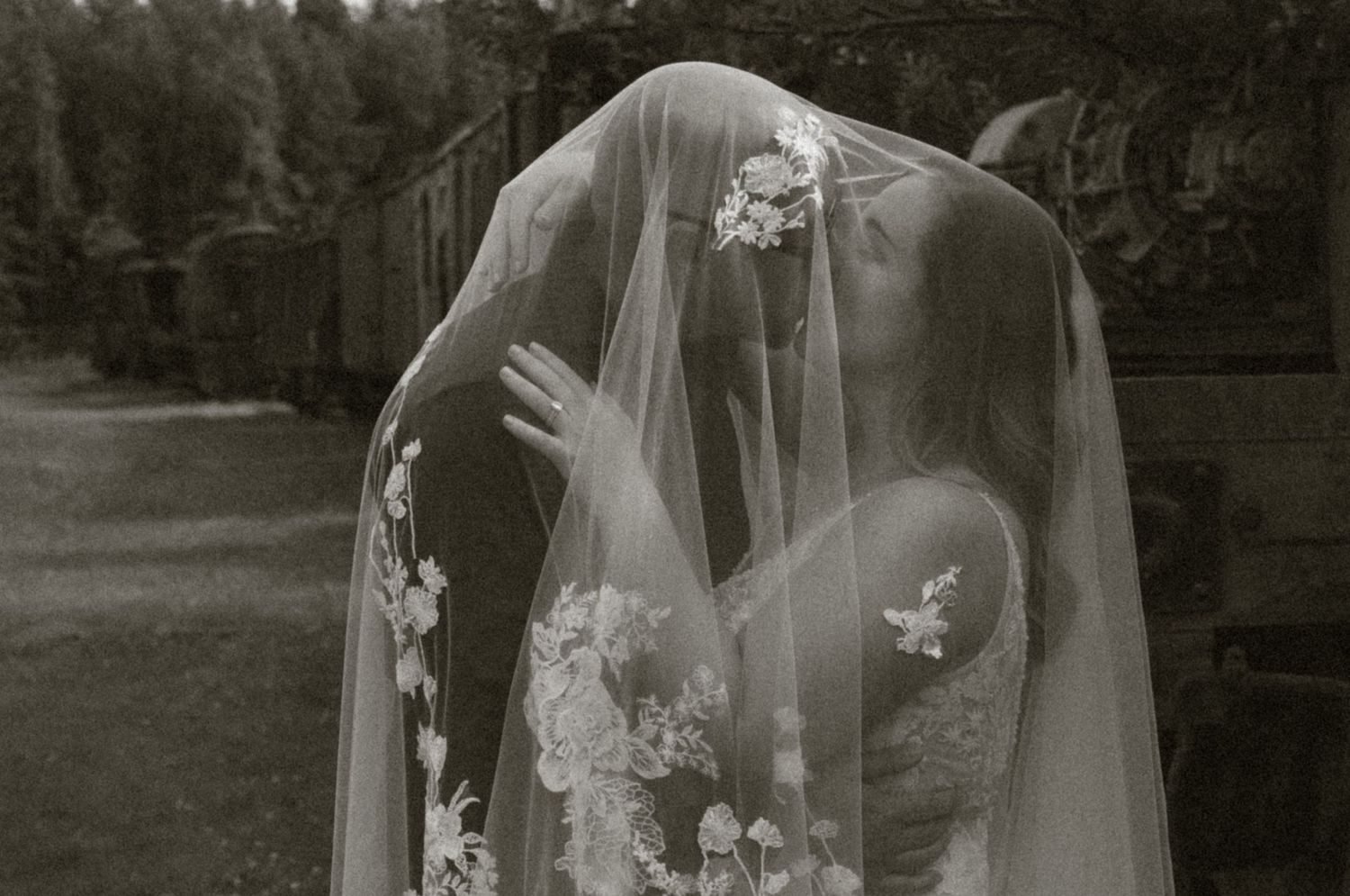 Black and white wedding photo of couple sharing a kiss under floral veil outdoors.
