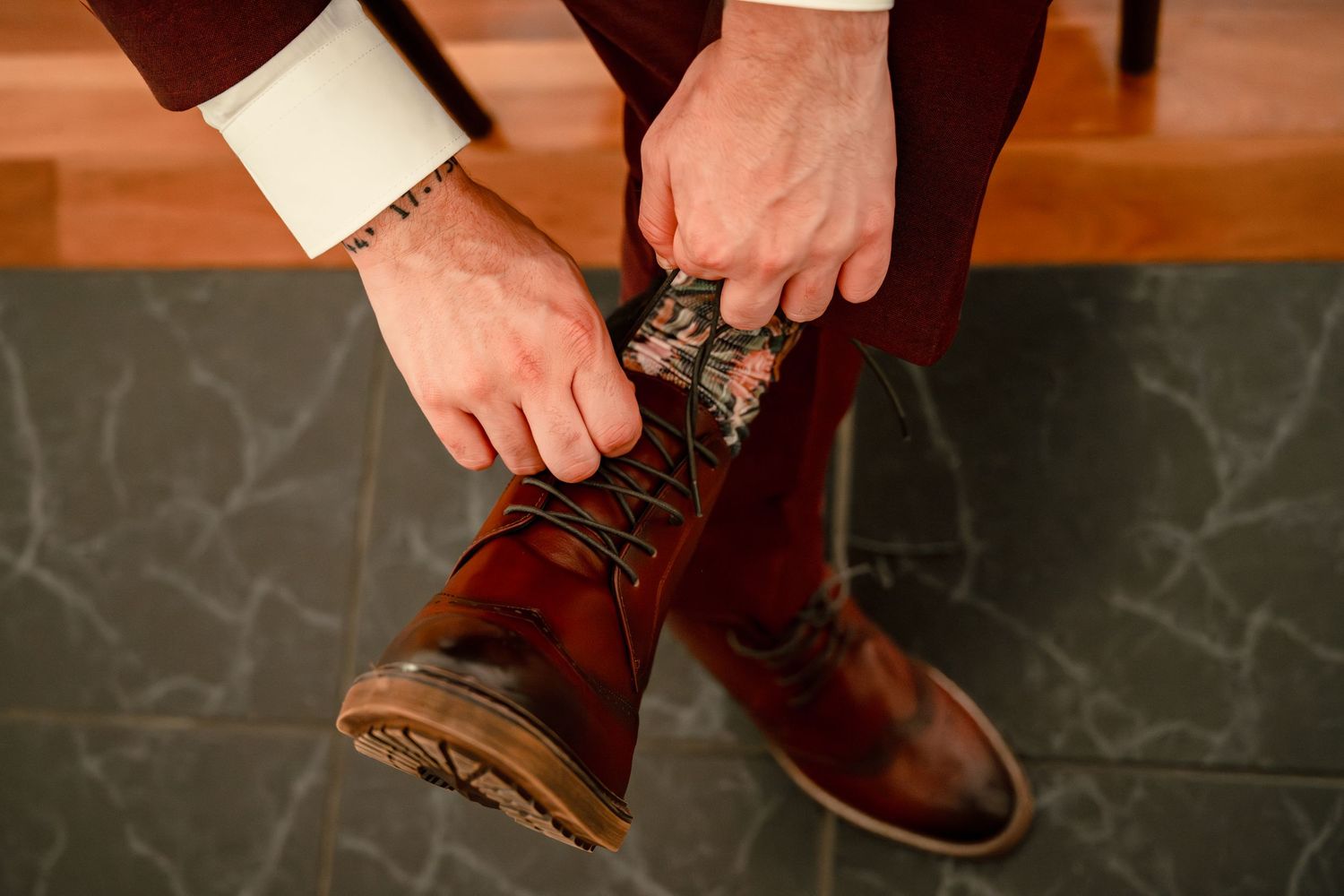Man putting on brown leather lace-up boots with patterned socks, wearing burgundy trousers and white dress shirt.