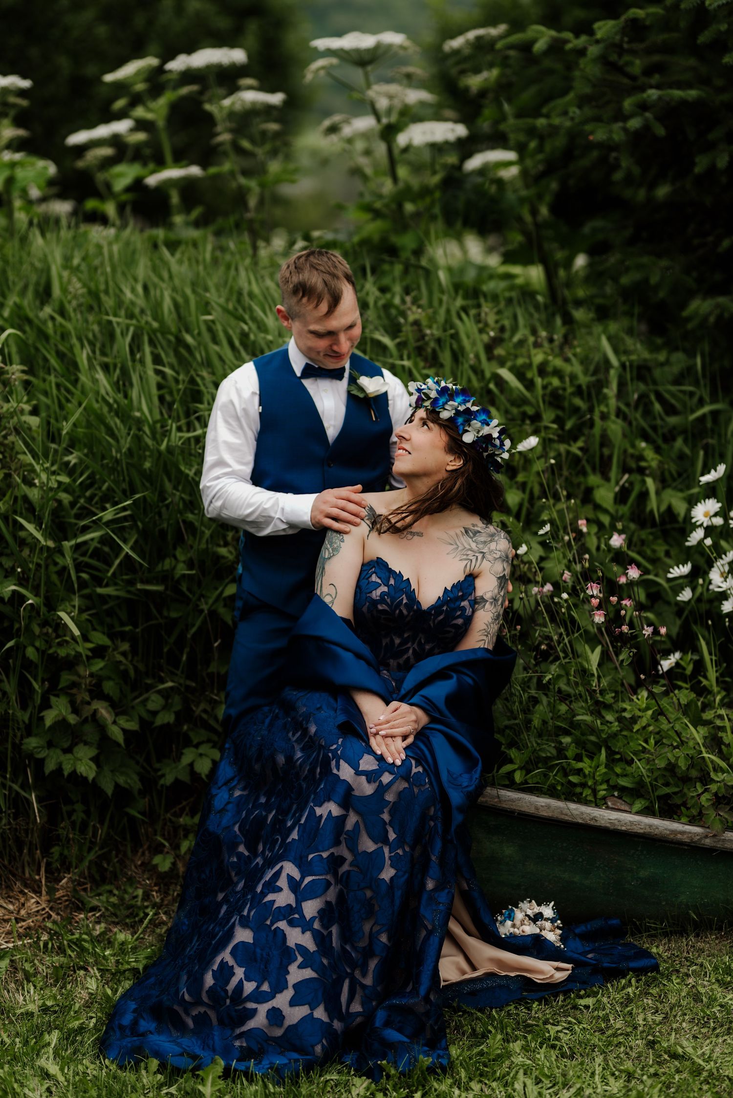 Bride in blue lace gown with floral crown sits in grass as groom in navy vest smiles down at her outdoors.
