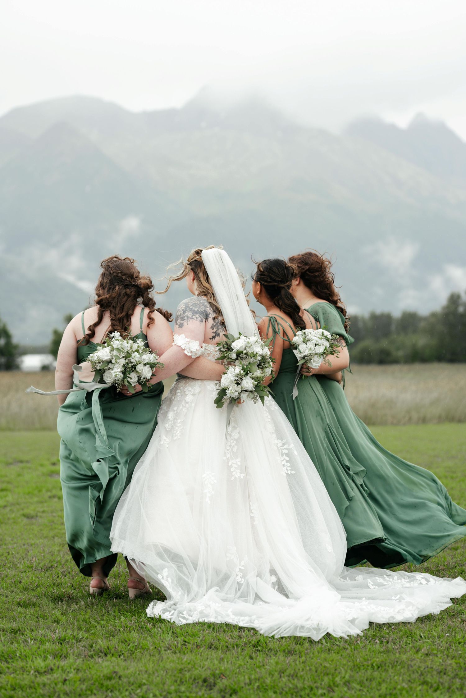 A bride and bridesmaids in sage green dresses walk together across a field with misty mountains in the background.