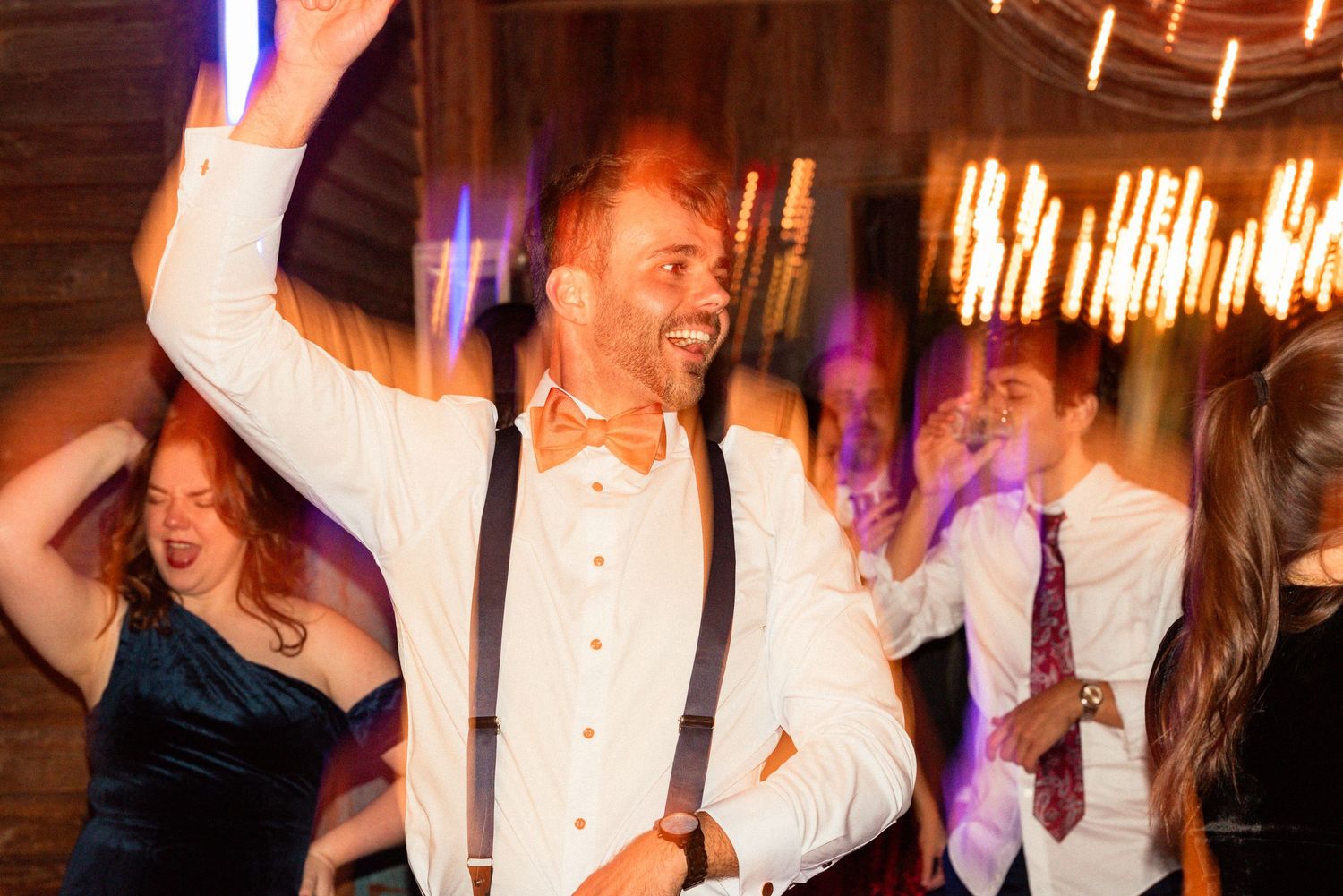Groom in white shirt and suspenders dancing joyfully at wedding reception with guests in warm ambient lighting.