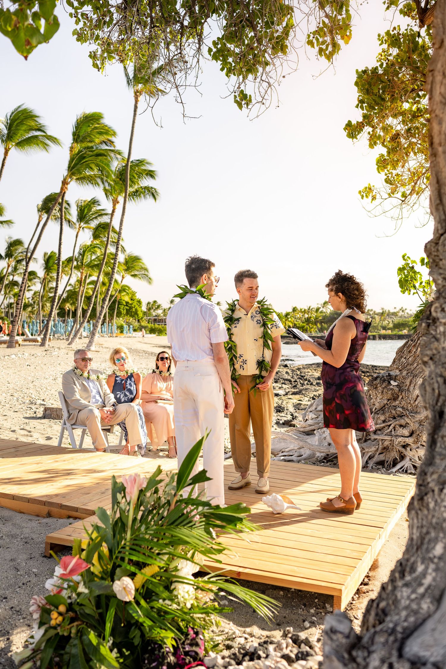 Same-sex couple exchanges vows on a wooden platform at a tropical beach wedding in Hawaii with palm trees.