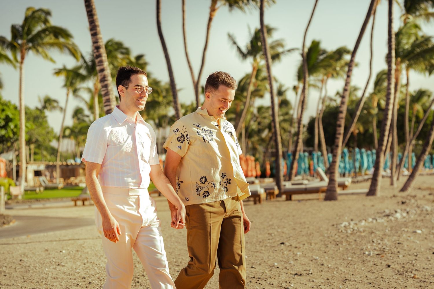 Two men walking on a tropical beach lined with palm trees, one in white outfit and other in floral shirt.