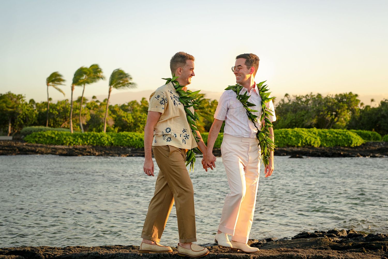 Two men wearing leis hold hands while walking along a Hawaiian shoreline at sunset.