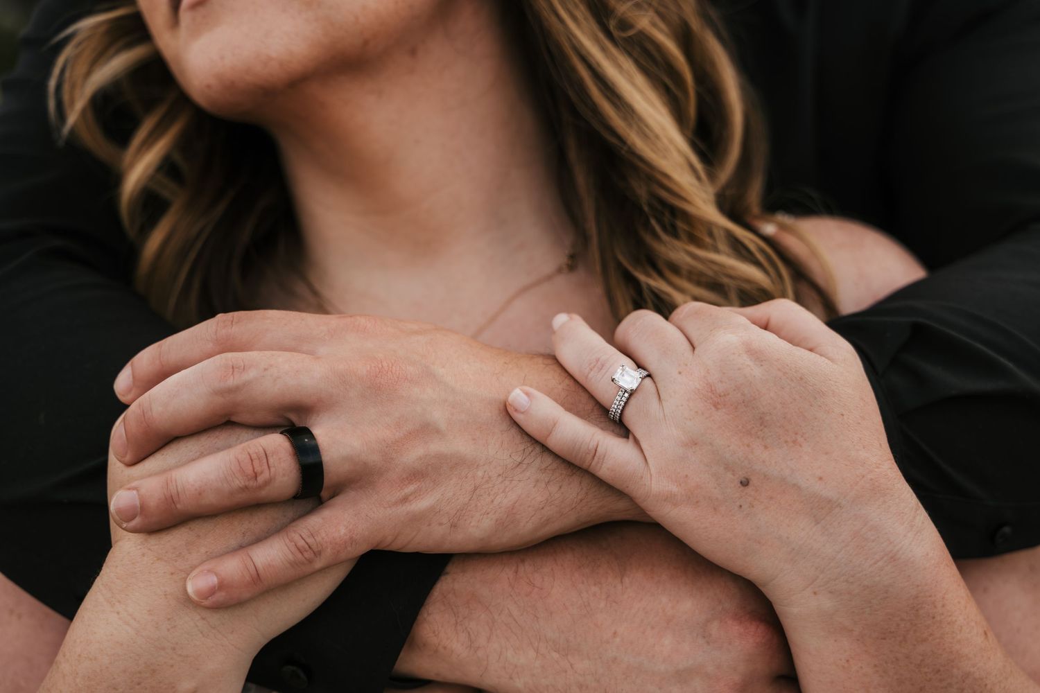 Couple embracing with hands crossed showing wedding rings, one black silicone band and one diamond engagement ring.