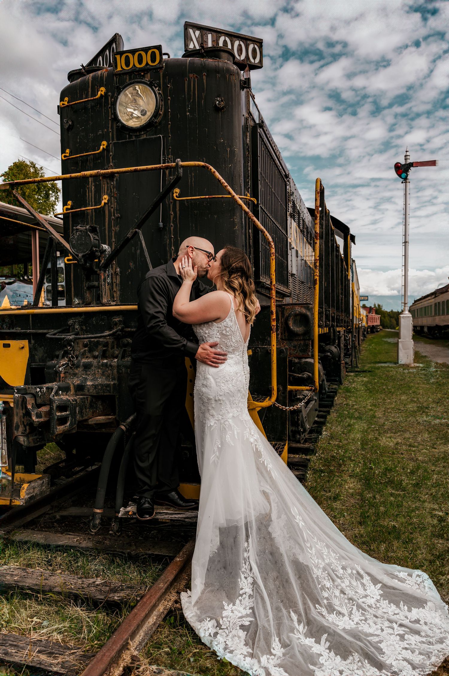 Bride and groom kissing in front of vintage black locomotive X1000 train, bride in lace mermaid gown with long train.