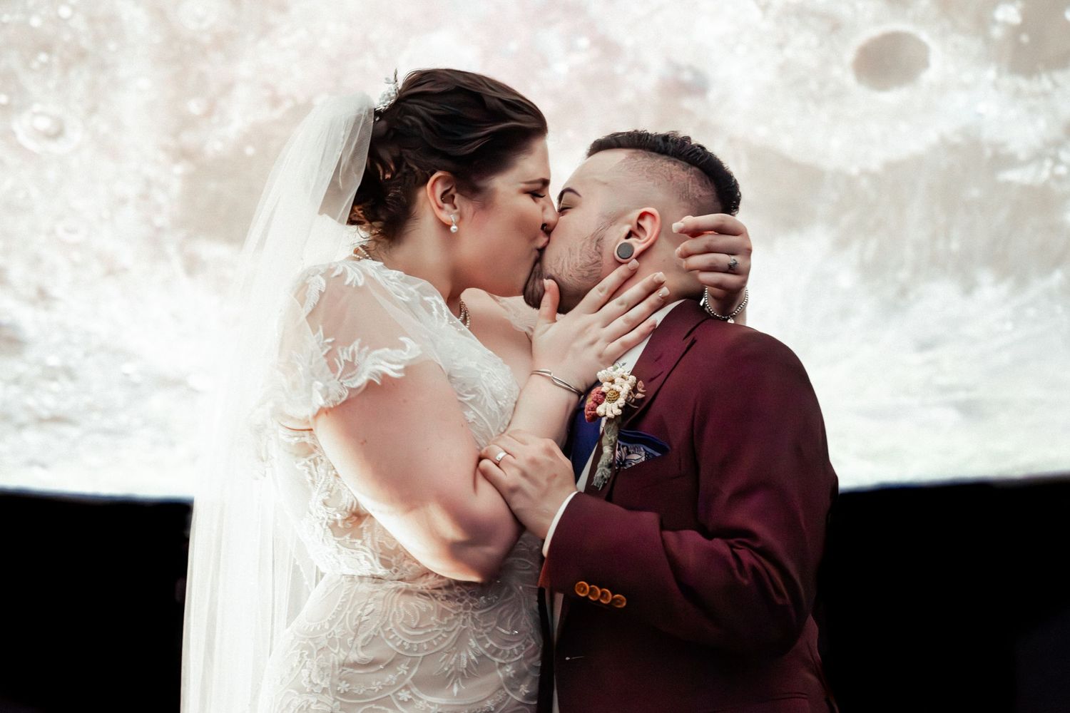 Bride and groom share first kiss at wedding ceremony with moon projection backdrop, bride in lace gown and groom in burgundy suit.
