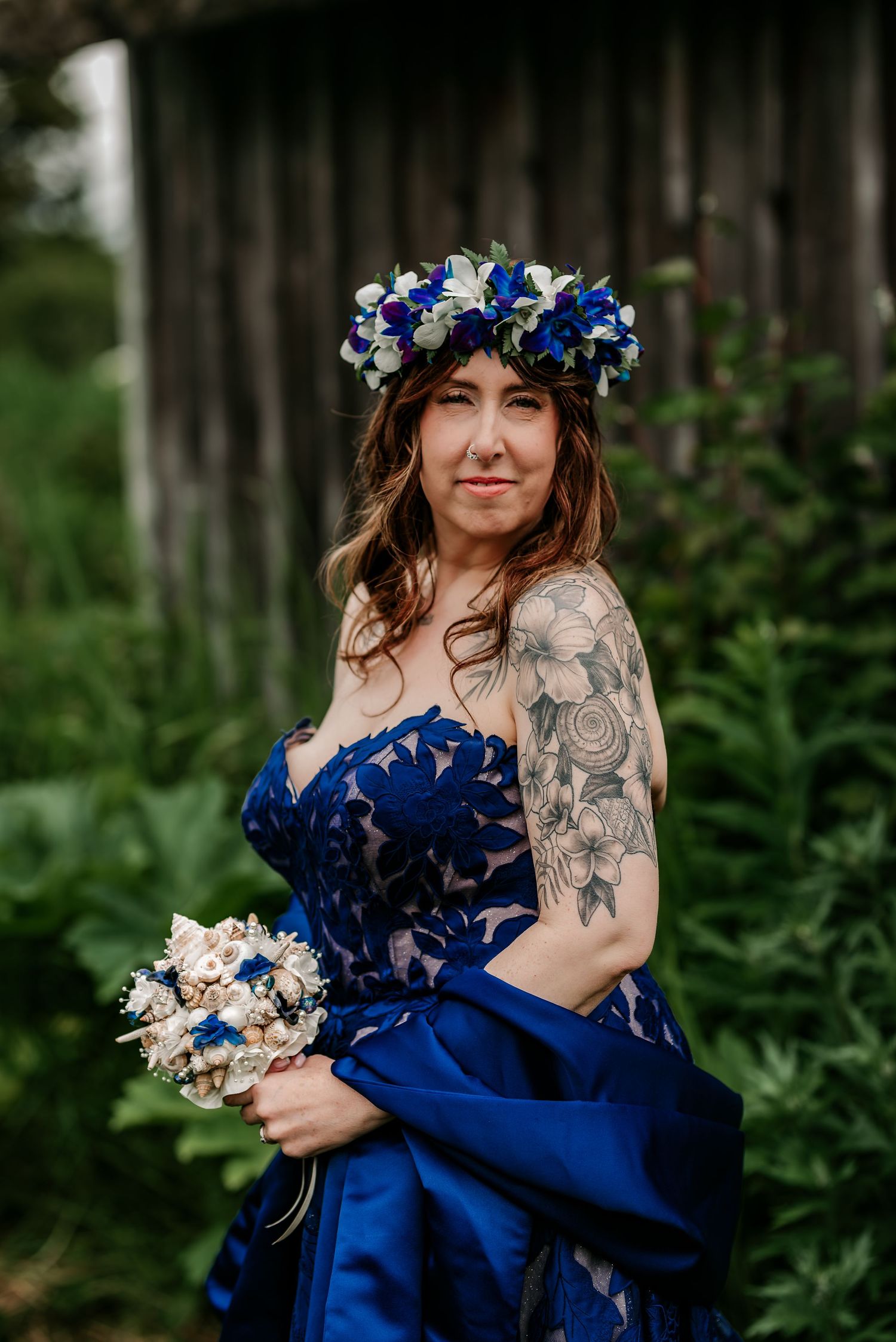 Bride in blue floral gown with flower crown and bouquet, showcasing tattoos, posing outdoors near rustic barn.