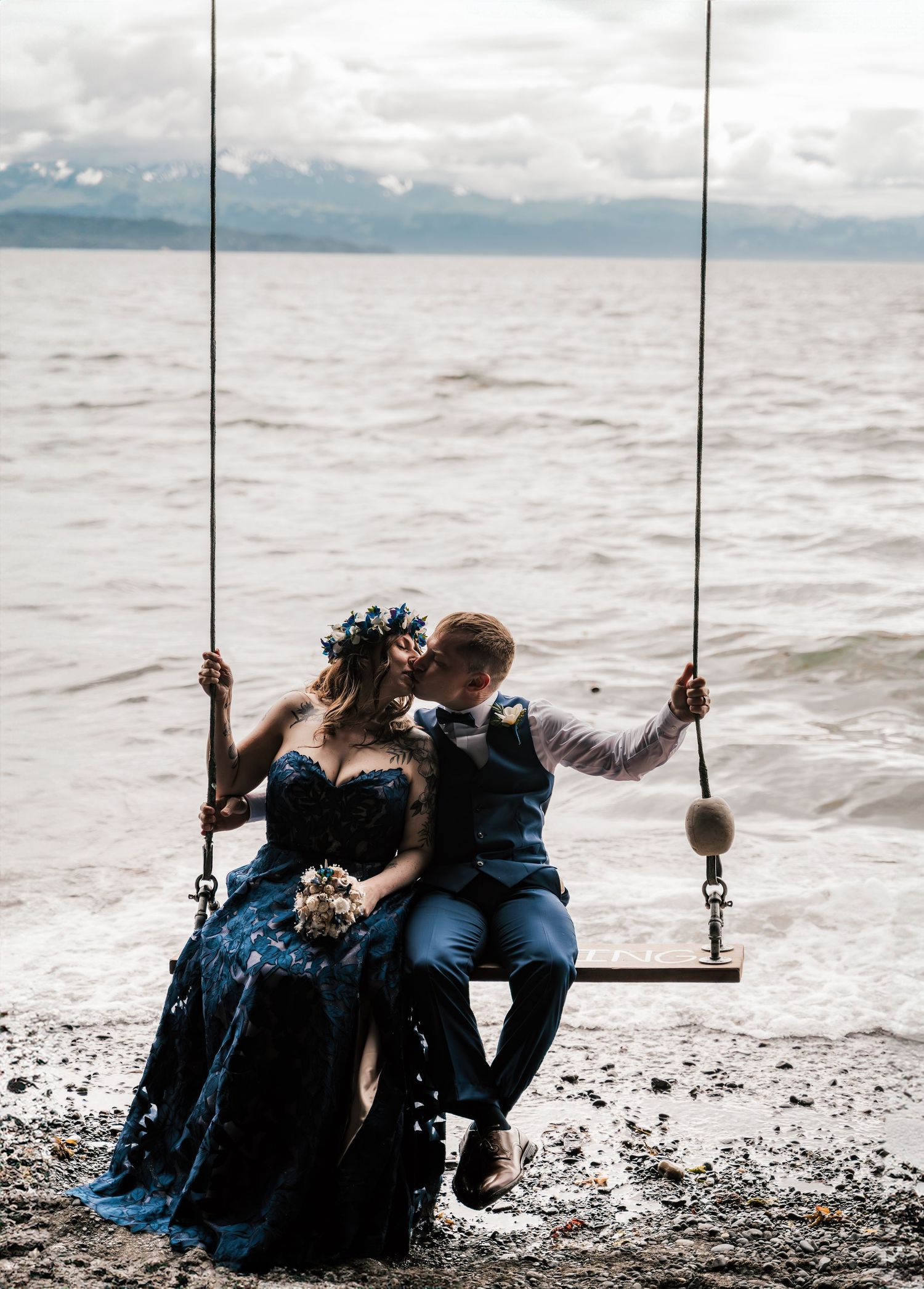 Couple in navy wedding attire sharing a romantic moment on a wooden swing In Homer Alaska with mountains in background.
