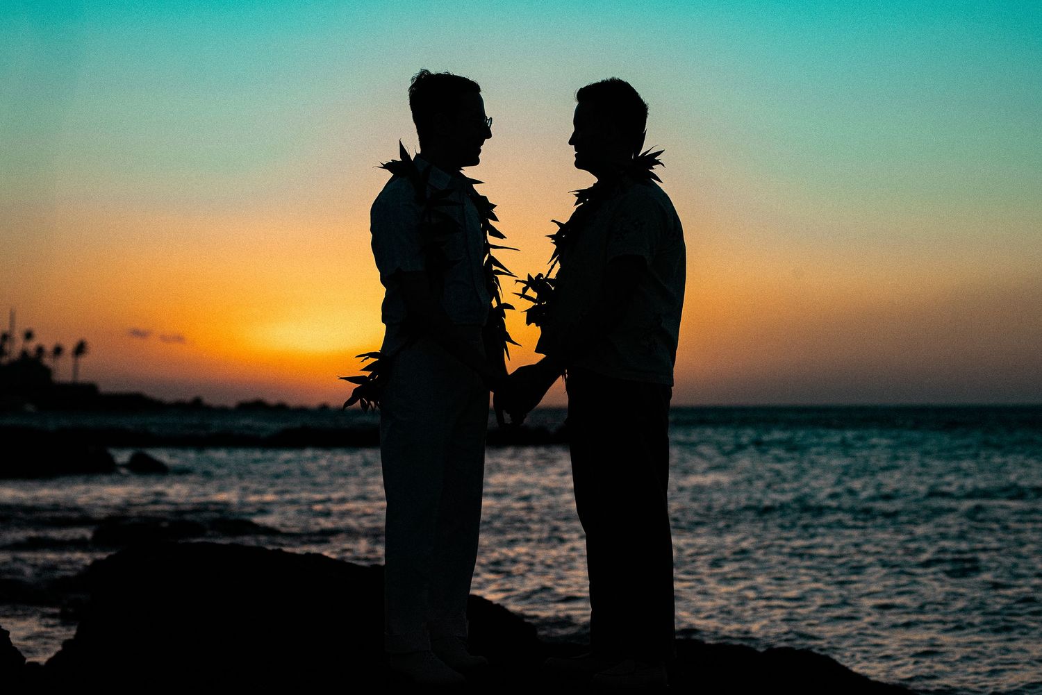 Silhouette of two grooms holding hands at sunset on a rocky Hawaiian beach during their wedding.