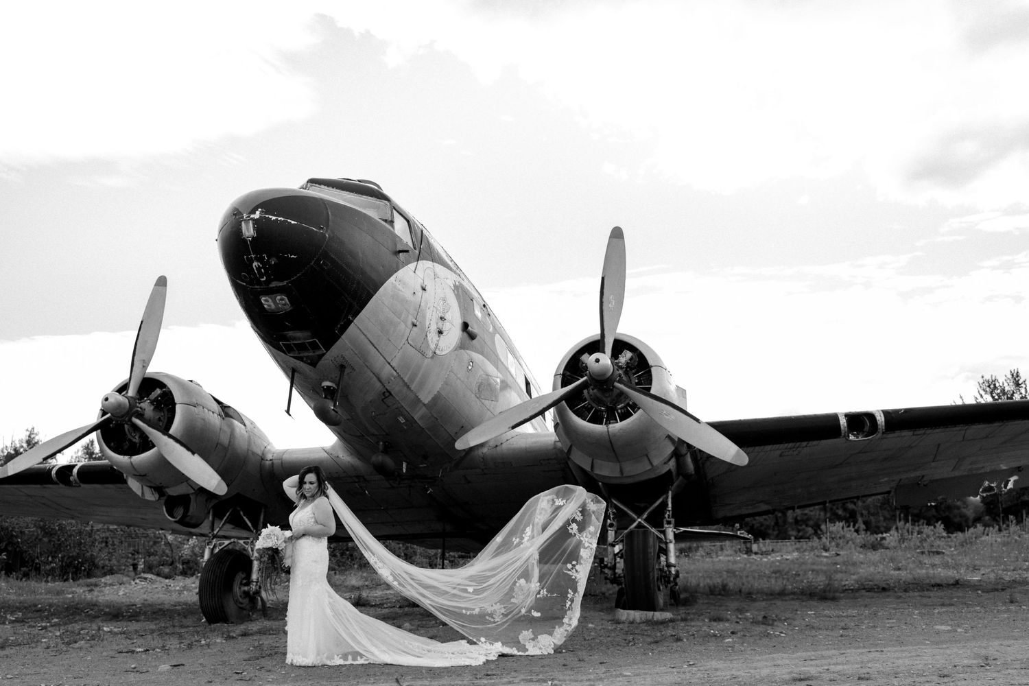 Bride in white gown poses before vintage twin-engine propeller aircraft in stunning black and white wedding photo palmer alaska