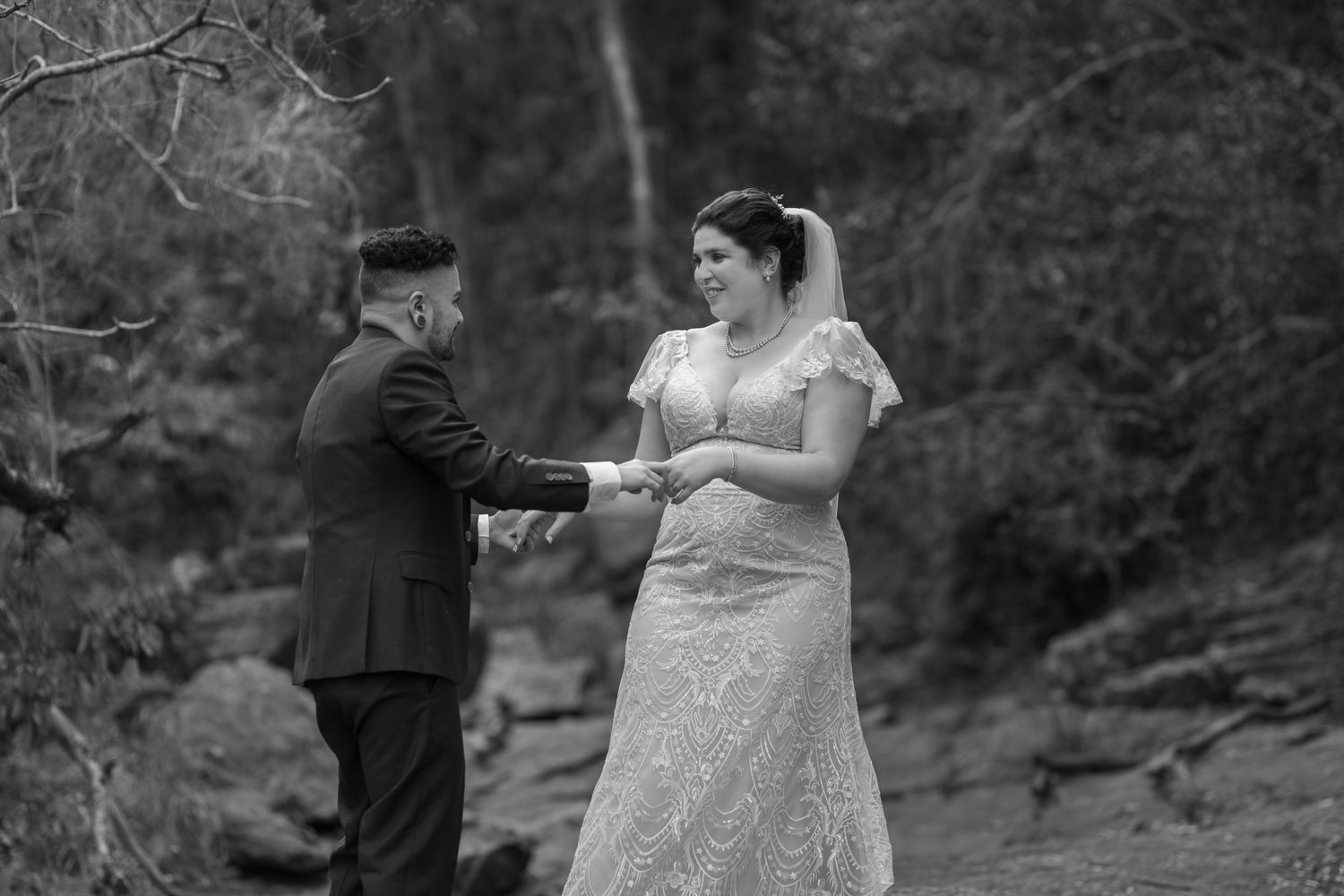 Black and white photo of a bride and groom exchanging vows outdoors in a lush forest setting.