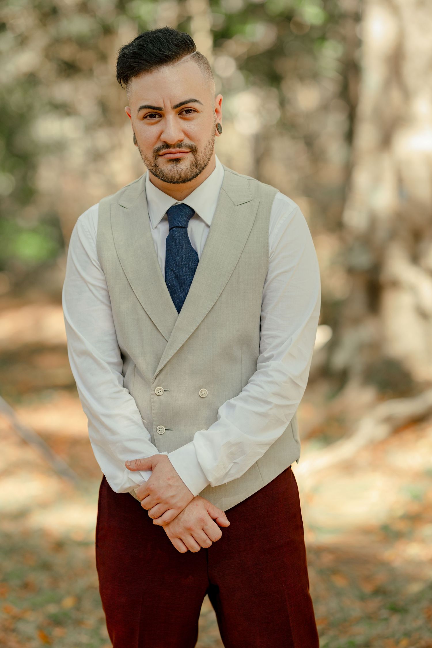A well-dressed man in a beige vest, white shirt, and blue tie with burgundy pants poses outdoors among trees.