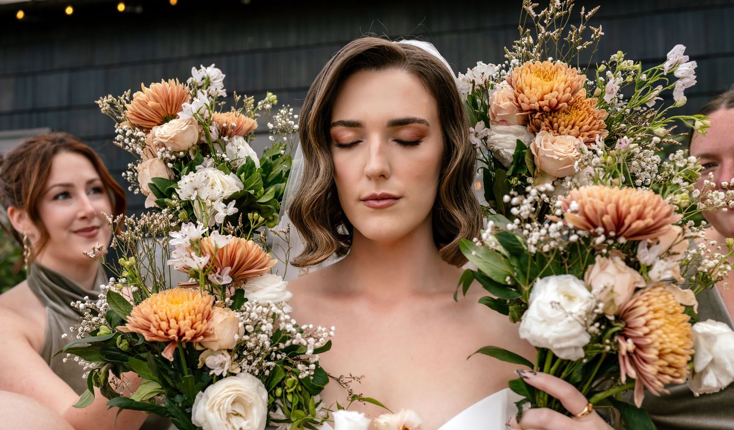 Bride holding a lush bouquet of peach dahlias and white roses, flanked by bridesmaids at an outdoor wedding.