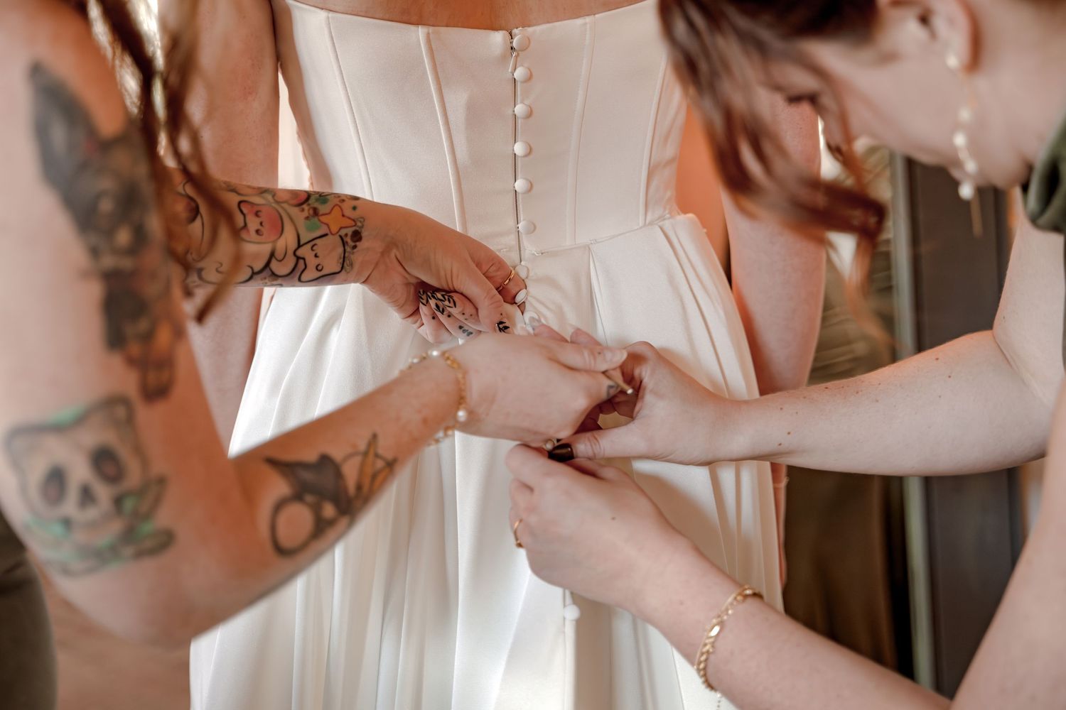 Bridesmaids helping bride fasten pearl bracelet on wrist, white corset wedding dress, tattooed arms visible.