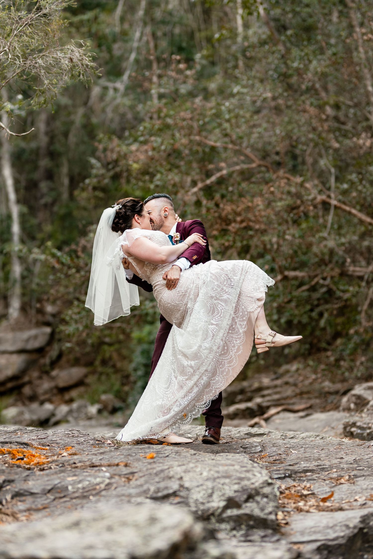 Groom lifts bride in lace wedding dress for a romantic kiss outdoors on rocky terrain surrounded by lush green trees.