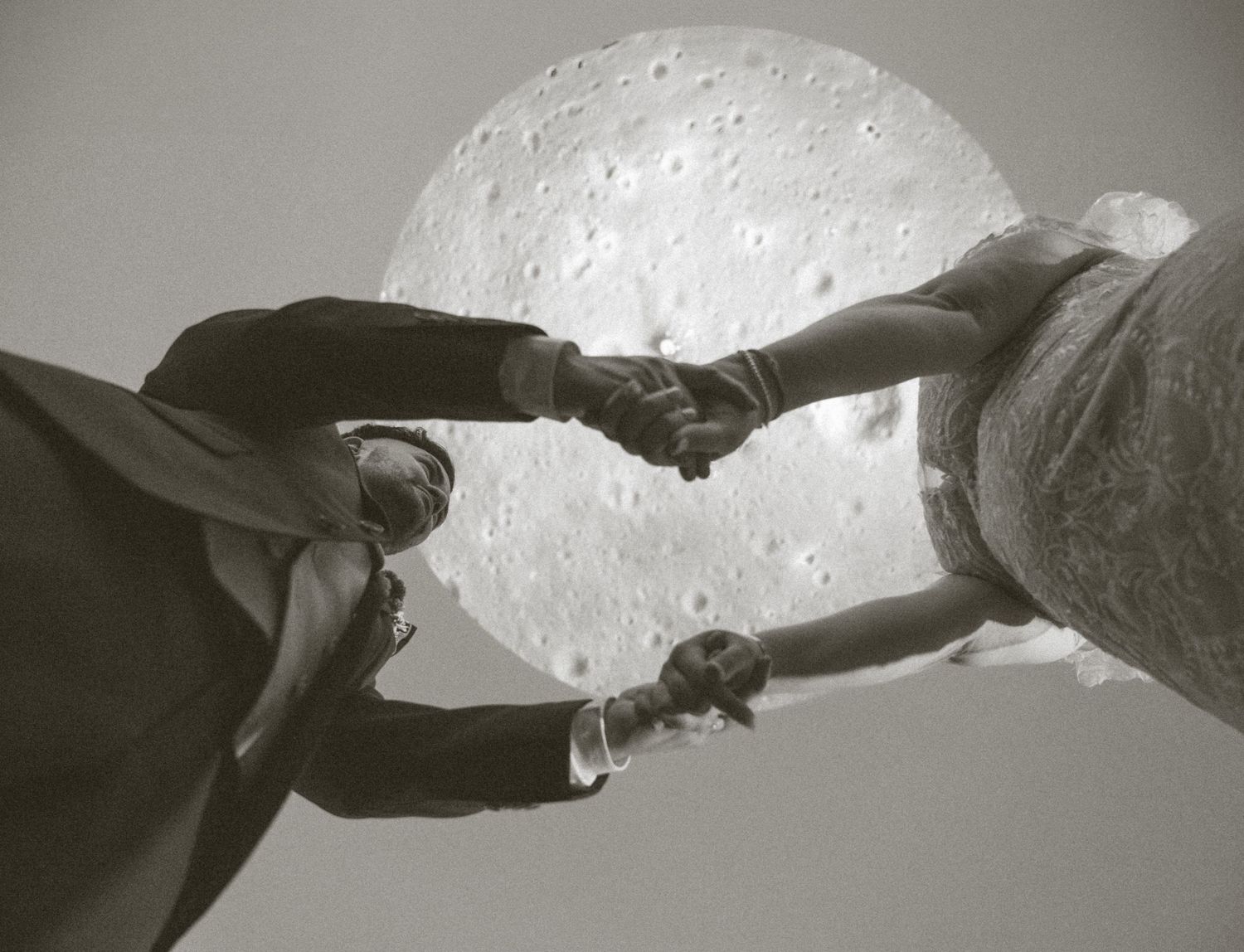 Two people holding hands in front of a large moon backdrop in a black and white artistic photograph.