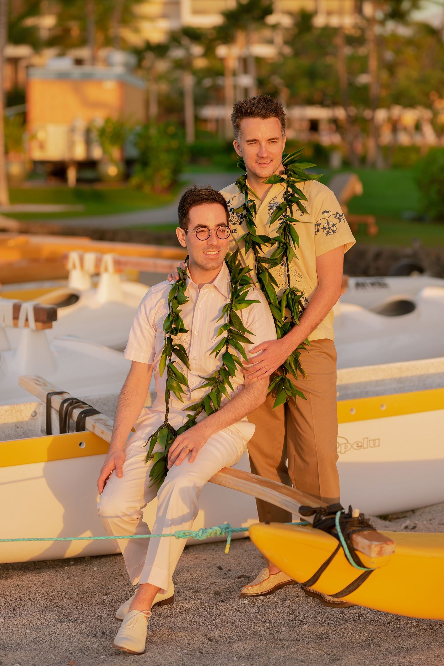 Two men wearing Hawaiian leis pose near outrigger canoes on a beach at golden hour.