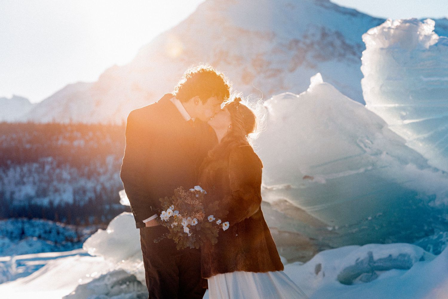 Bride and groom sharing a romantic kiss surrounded by snow and ice with golden sunlight glowing behind them.