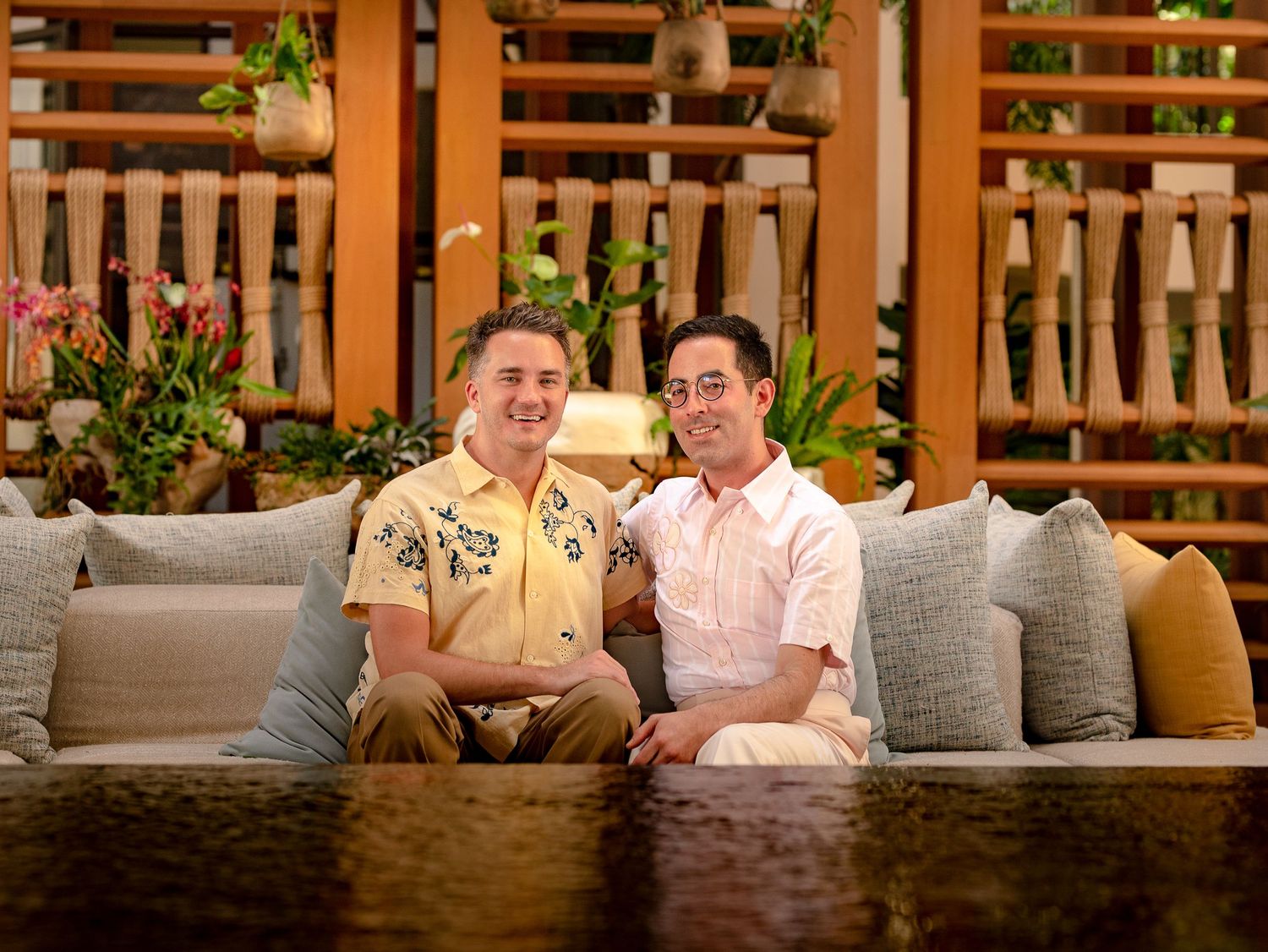 Two smiling men sit together on a cozy outdoor sofa surrounded by tropical plants and wooden decor.