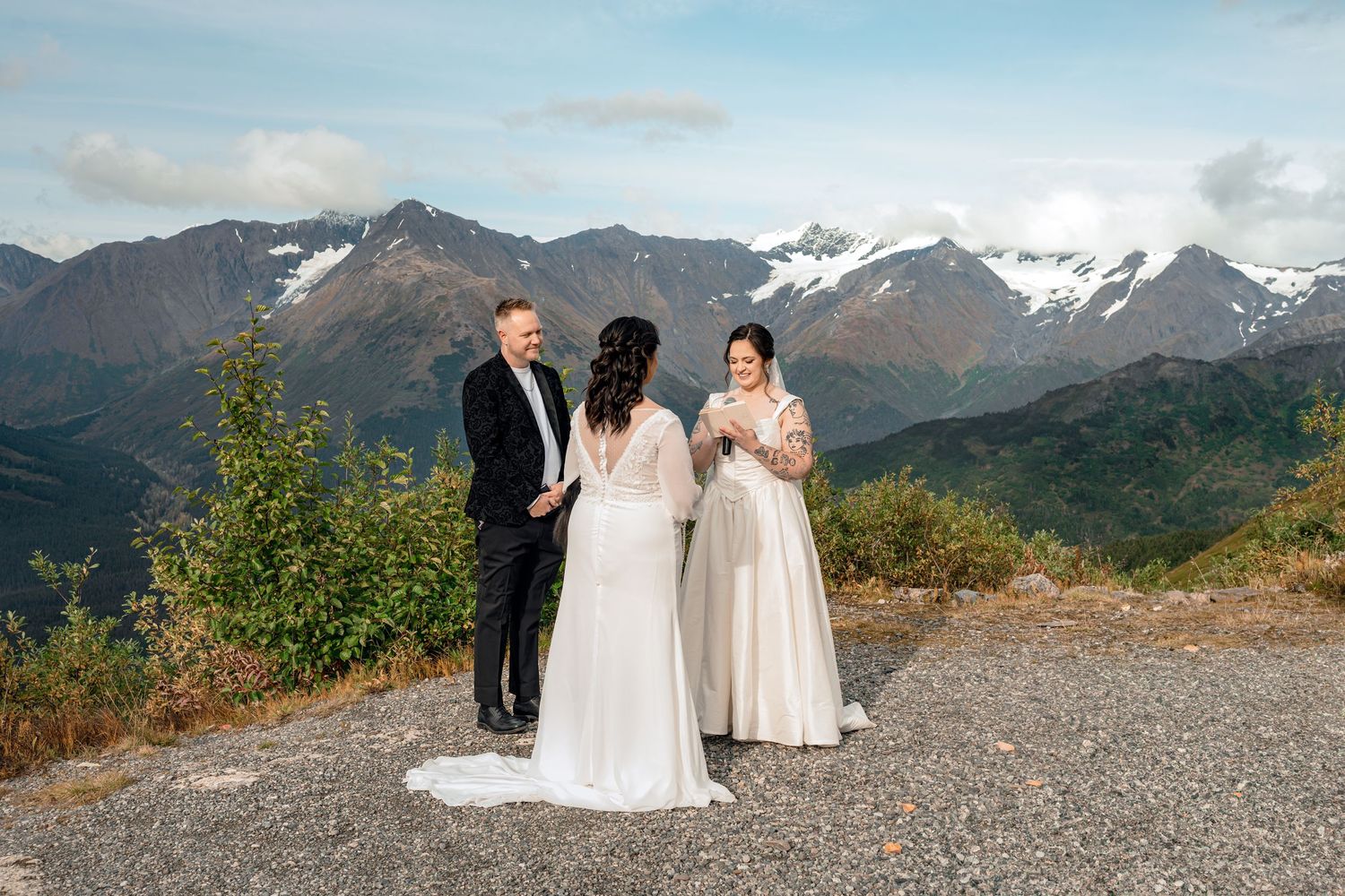 Couple exchanges vows with officiant on mountain overlook with snow-capped peaks in Alaska backdrop.