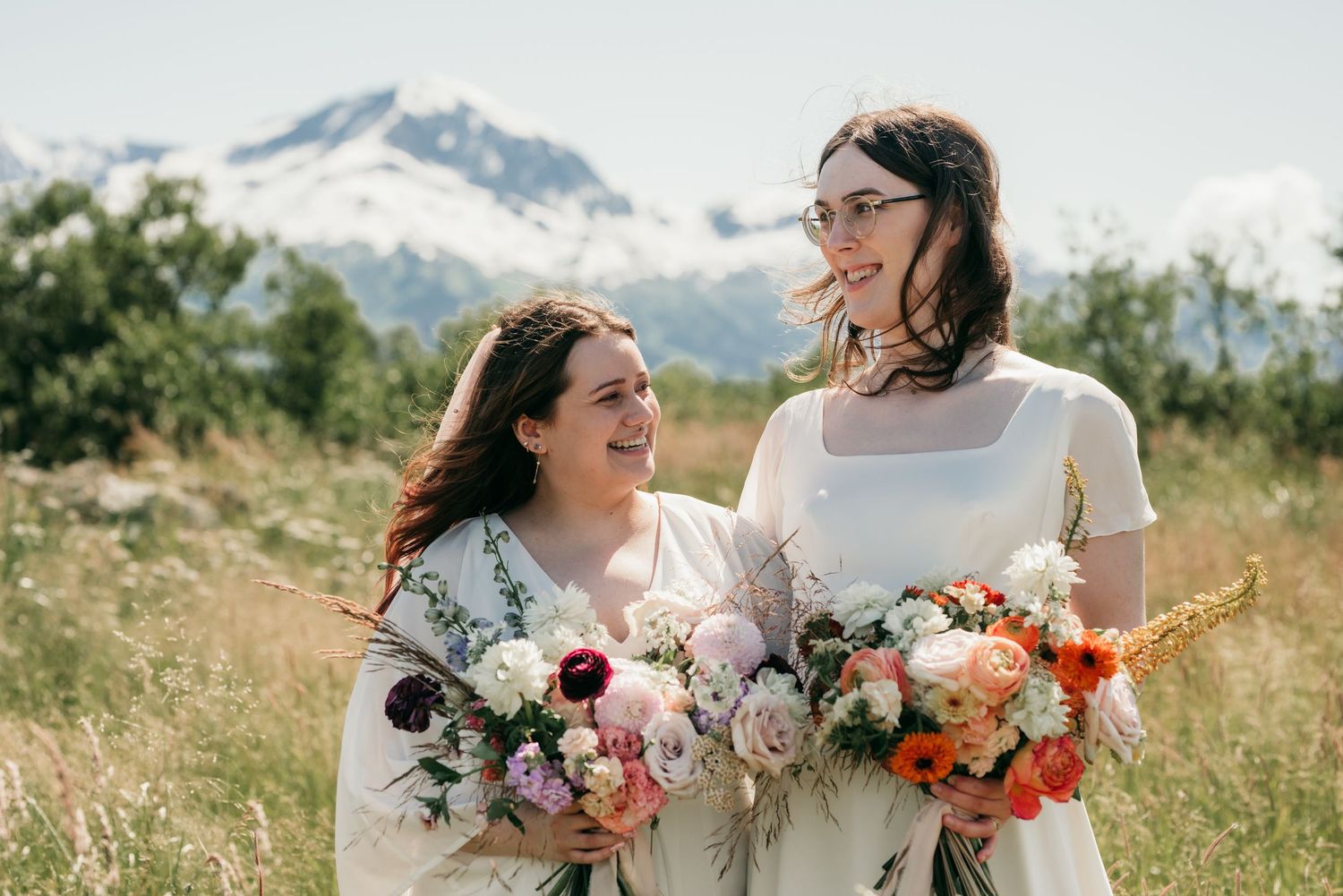 Two people in wedding attire hold colorful bouquets of flowers with snow-capped mountains in the background.