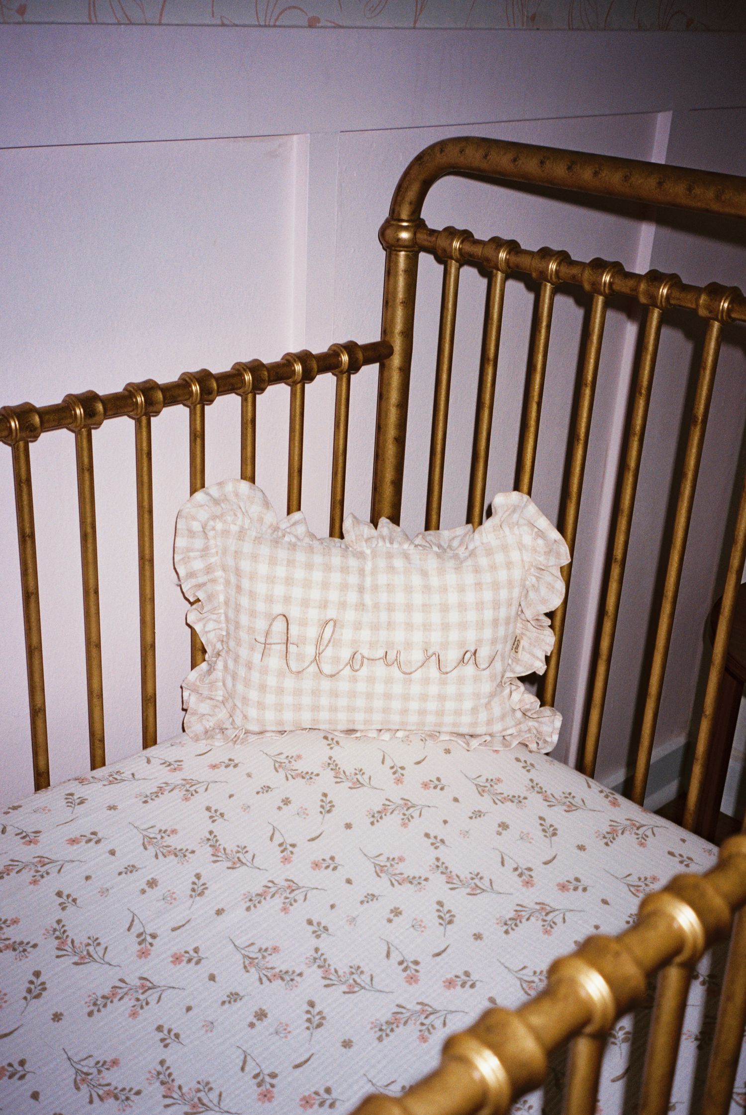Gold metal crib with floral bedding and a ruffled gingham pillow embroidered with the name Aloura in a nursery.