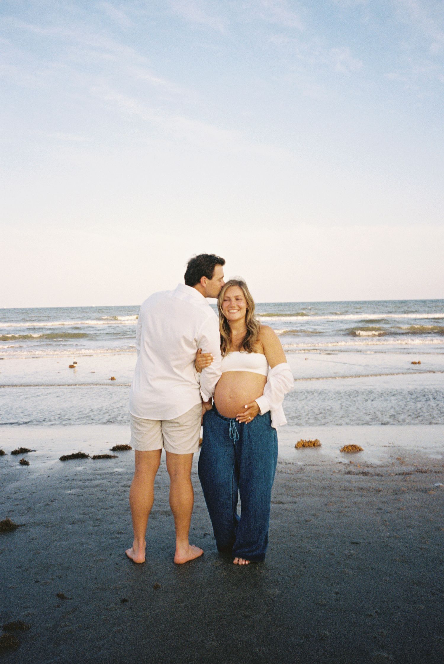 Pregnant couple embracing on beach at sunset, woman in white top and jeans cradling baby bump, man kissing her cheek.