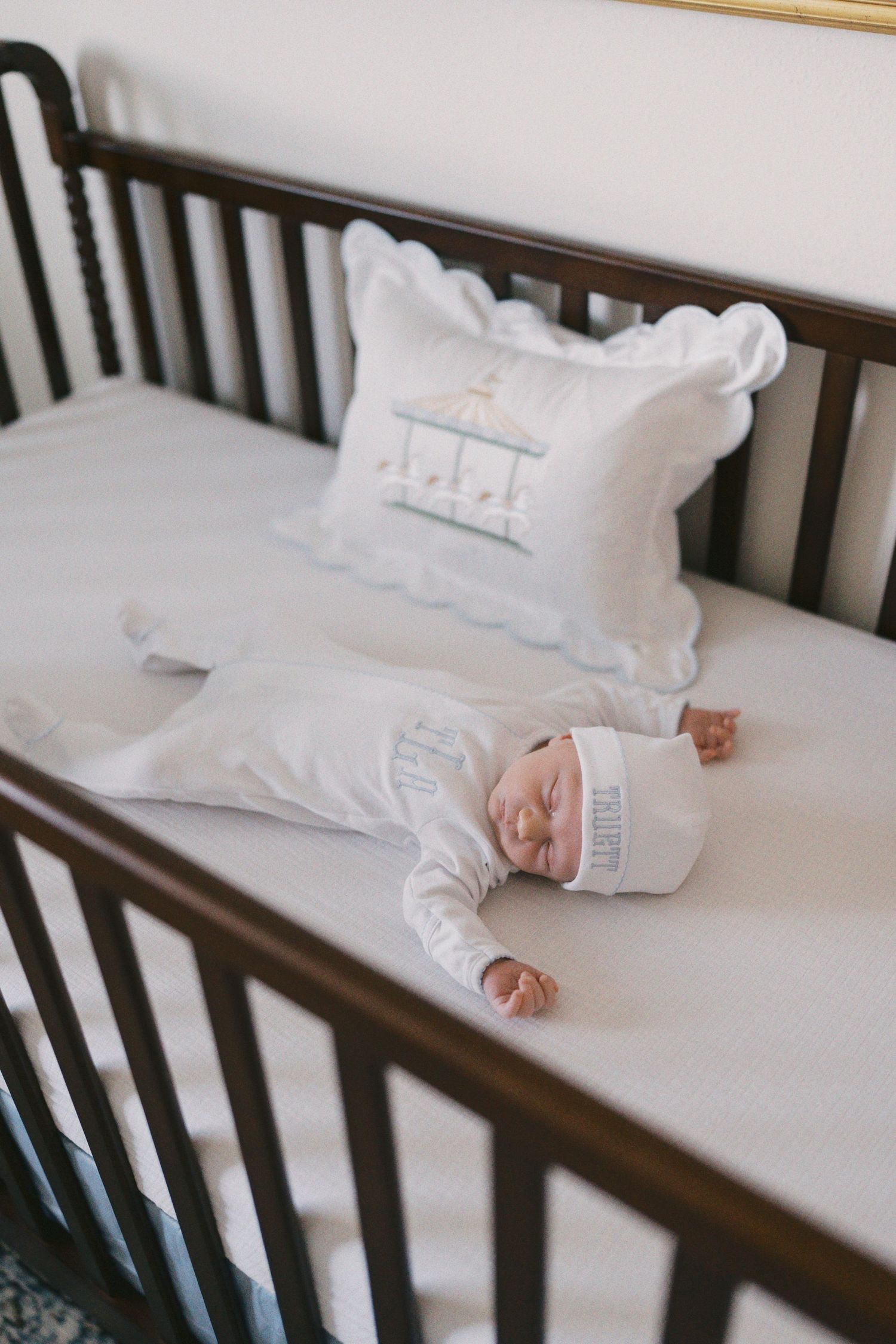 Newborn baby sleeping peacefully in a dark wood crib wearing white outfit and hat with decorative pillow.