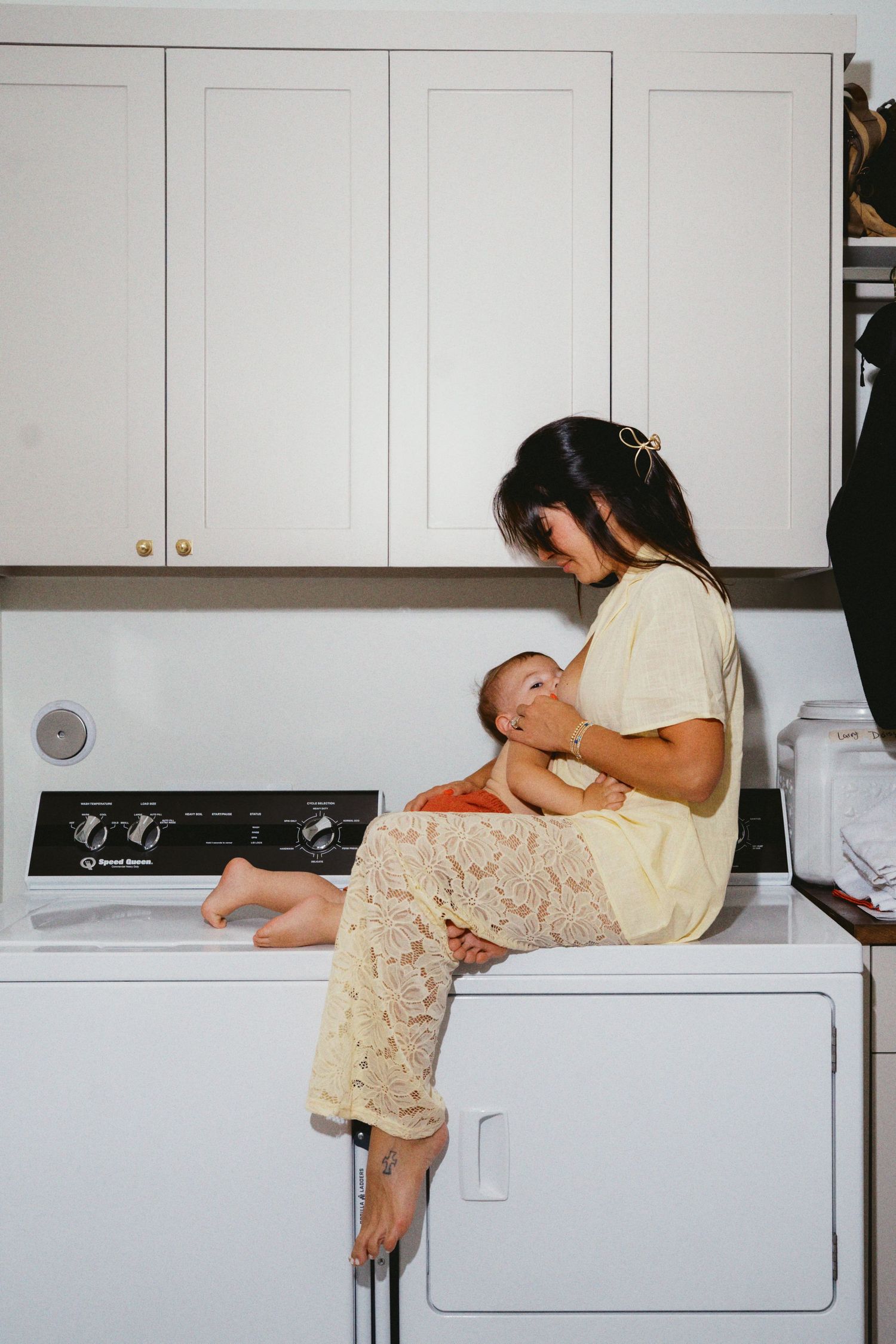 Mother breastfeeding newborn baby while sitting on washing machine in laundry room.