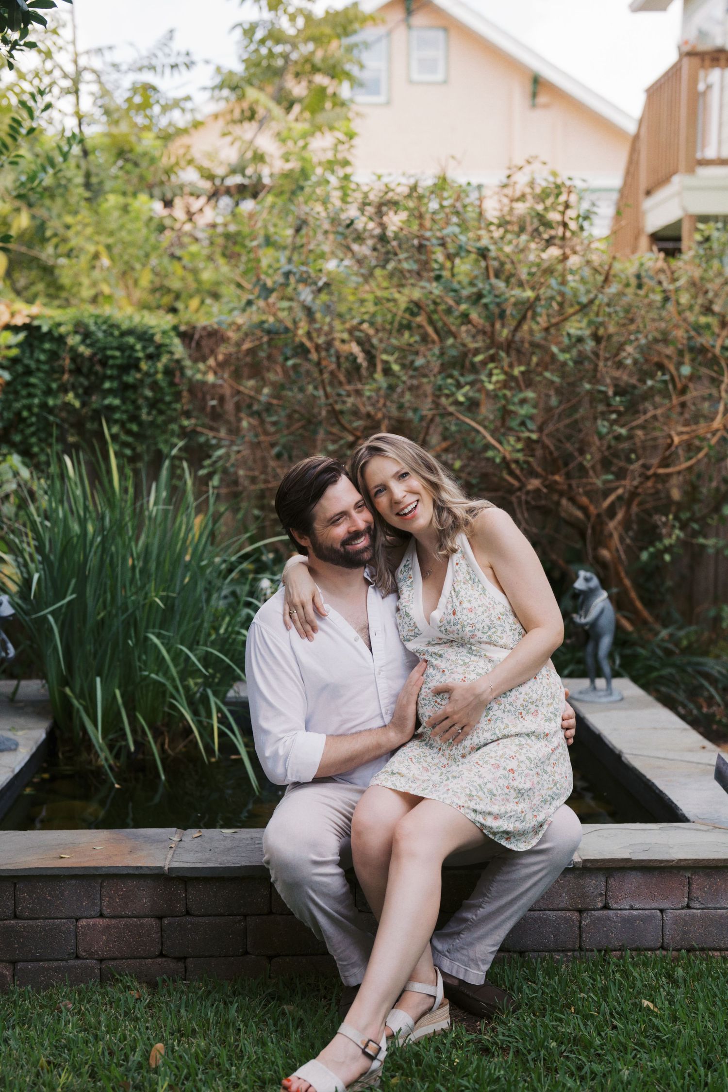 Happy couple sitting by a garden pond, woman in floral dress cradling baby bump, man in white shirt embracing her.