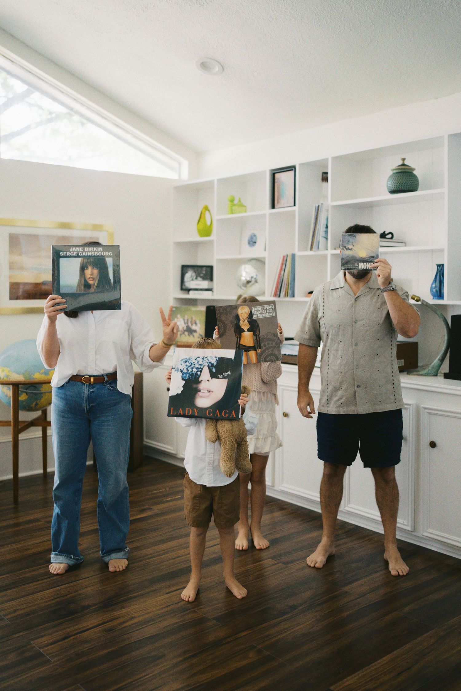 Family of three holding vinyl records over their faces in a modern living room with built-in shelves.
