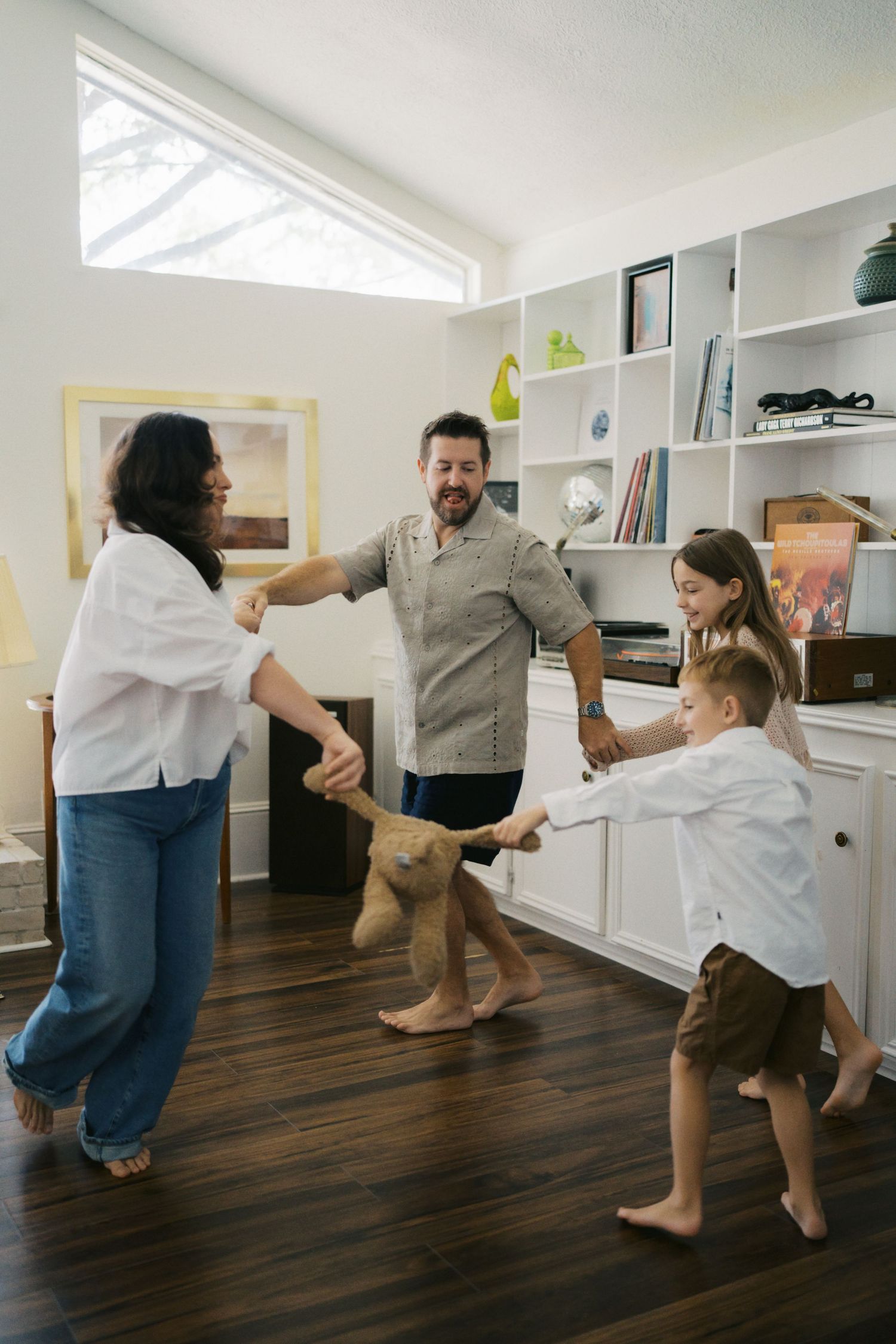 Family of four playing tug-of-war with a teddy bear in a modern living room with hardwood floors.
