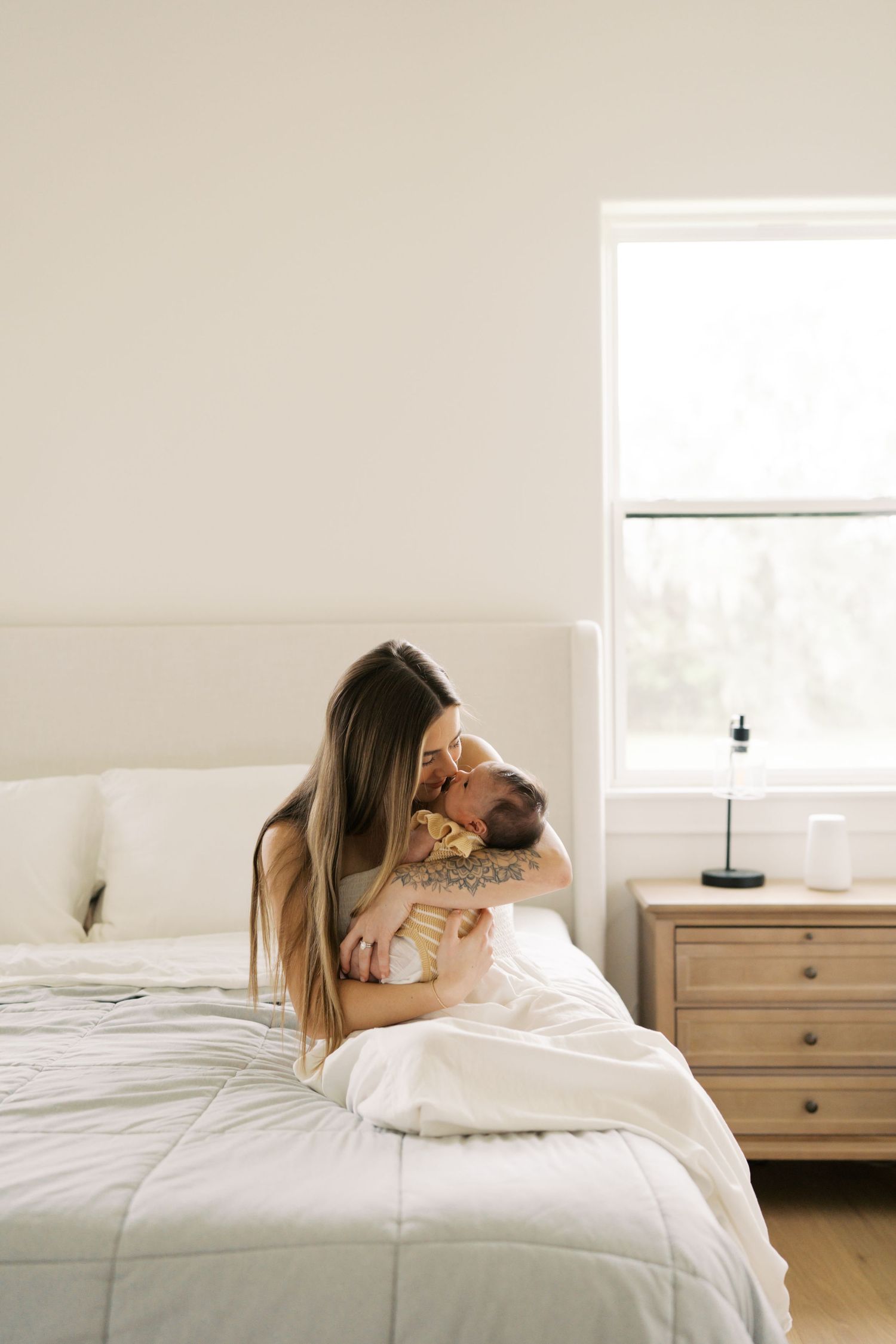 A mother with long hair sits on a white bed, gently kissing her newborn baby near a bright window.