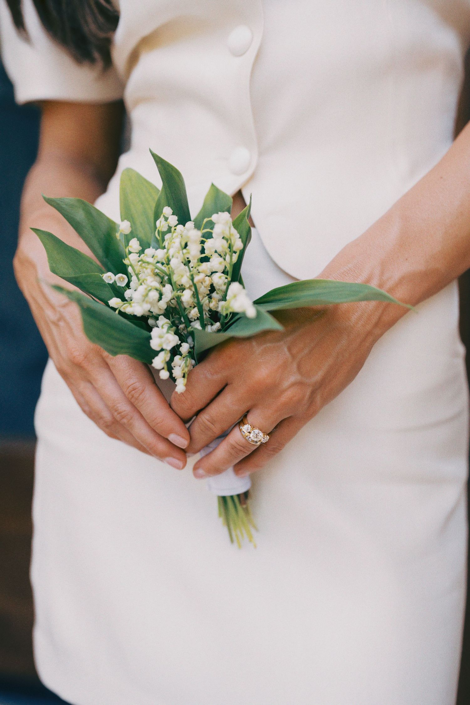Bride holding a small lily of the valley bouquet with green leaves, wearing a white dress and diamond engagement ring.
