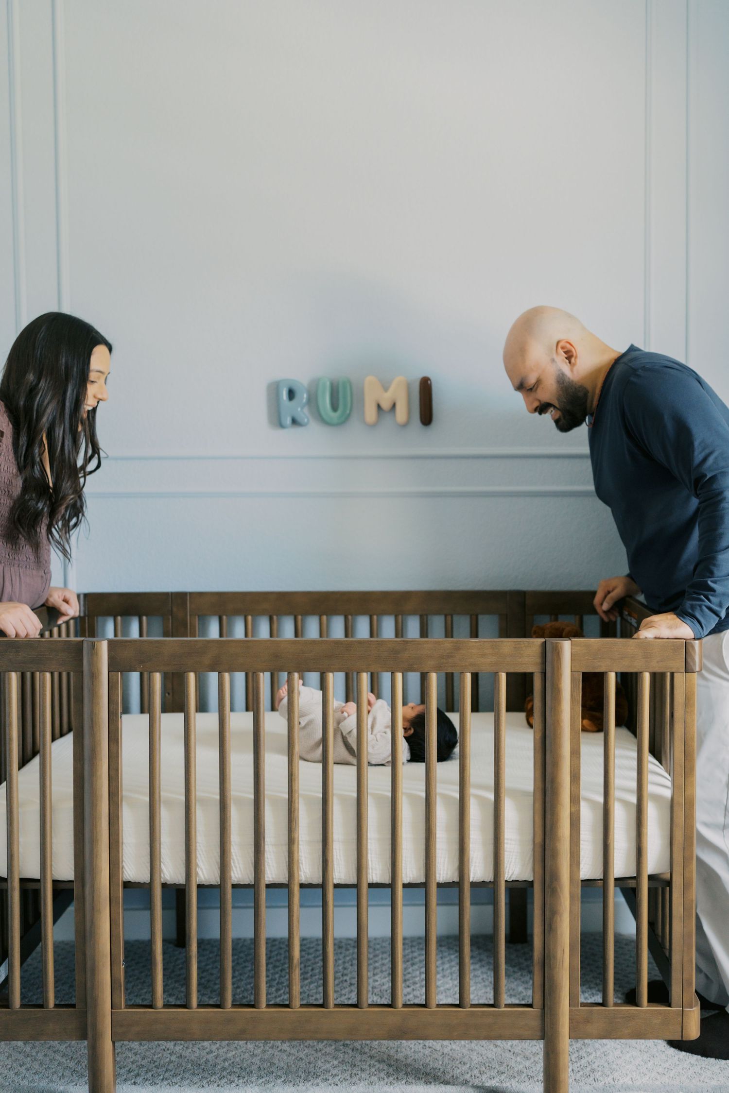 Parents leaning over a wooden crib with baby named Rumi, colorful name letters displayed on nursery wall.
