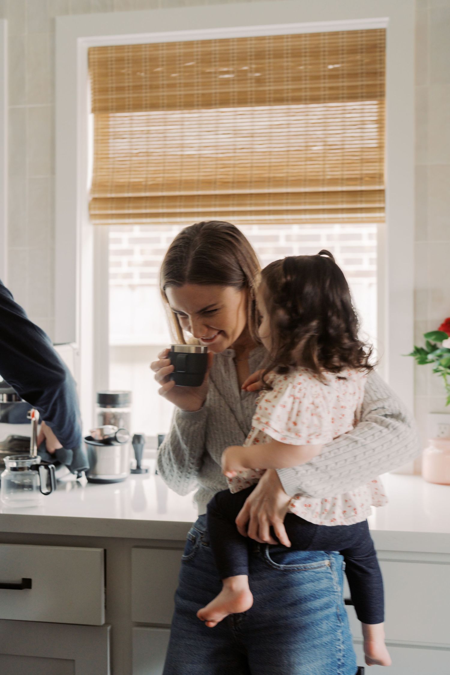 A young woman holding a young child in a kitchen with a window and blinds in the background.