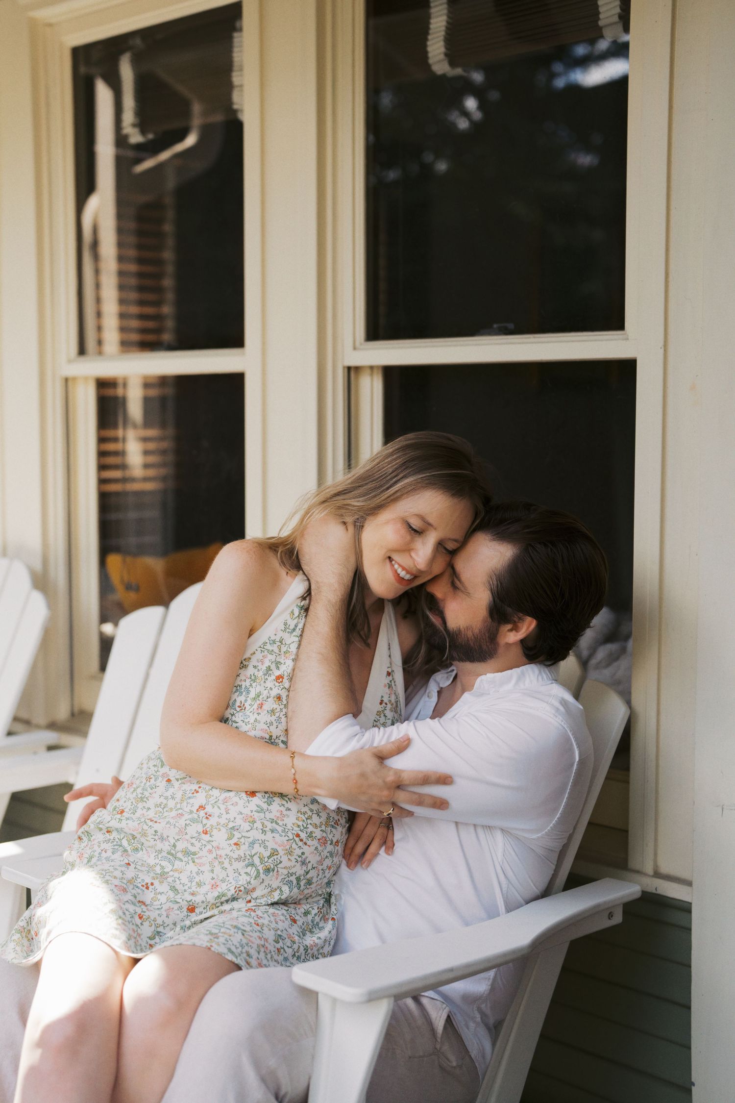 Couple sharing a tender moment on a porch, woman in floral dress sitting on man's lap in white Adirondack chair.