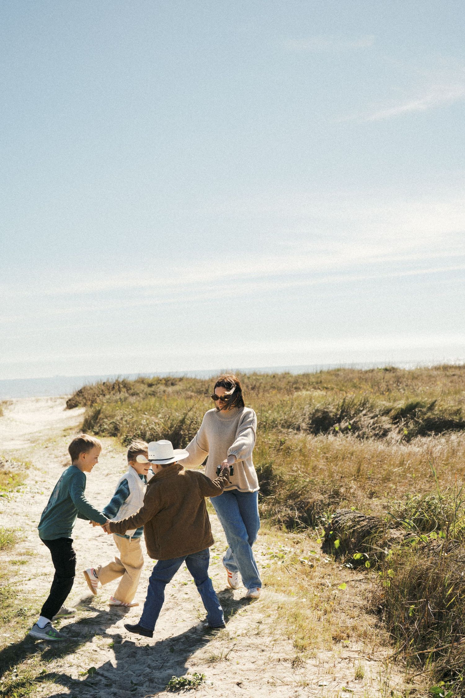 A family enjoys a sunny day walking together on a coastal trail with grassy dunes and blue sky in the background.