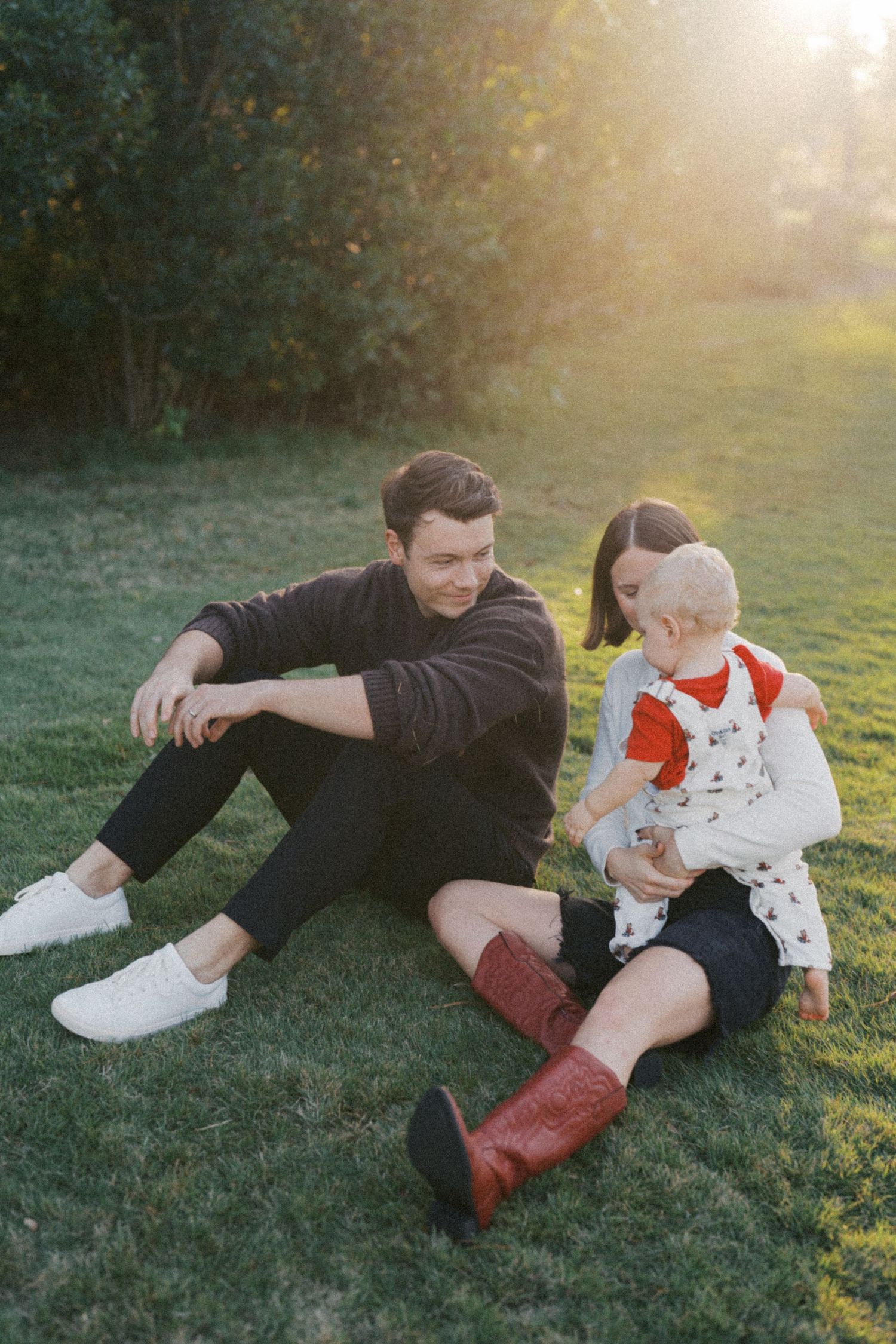 Two people sit together on grass during golden hour, with one wearing black and another in white with red boots.