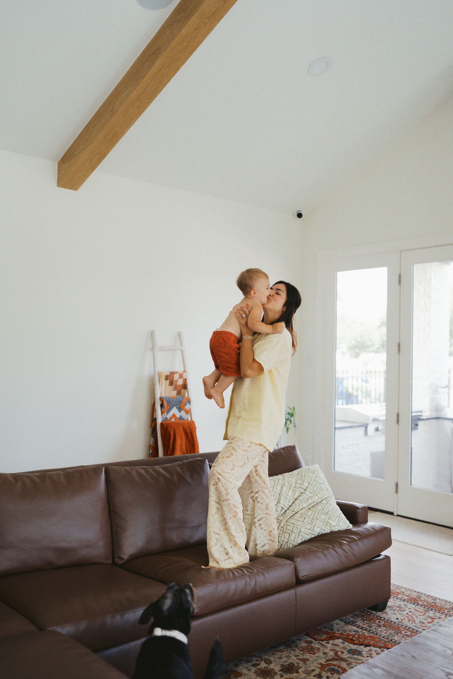 A mother lifts and kisses her baby while standing on a leather sofa in a modern white living room.