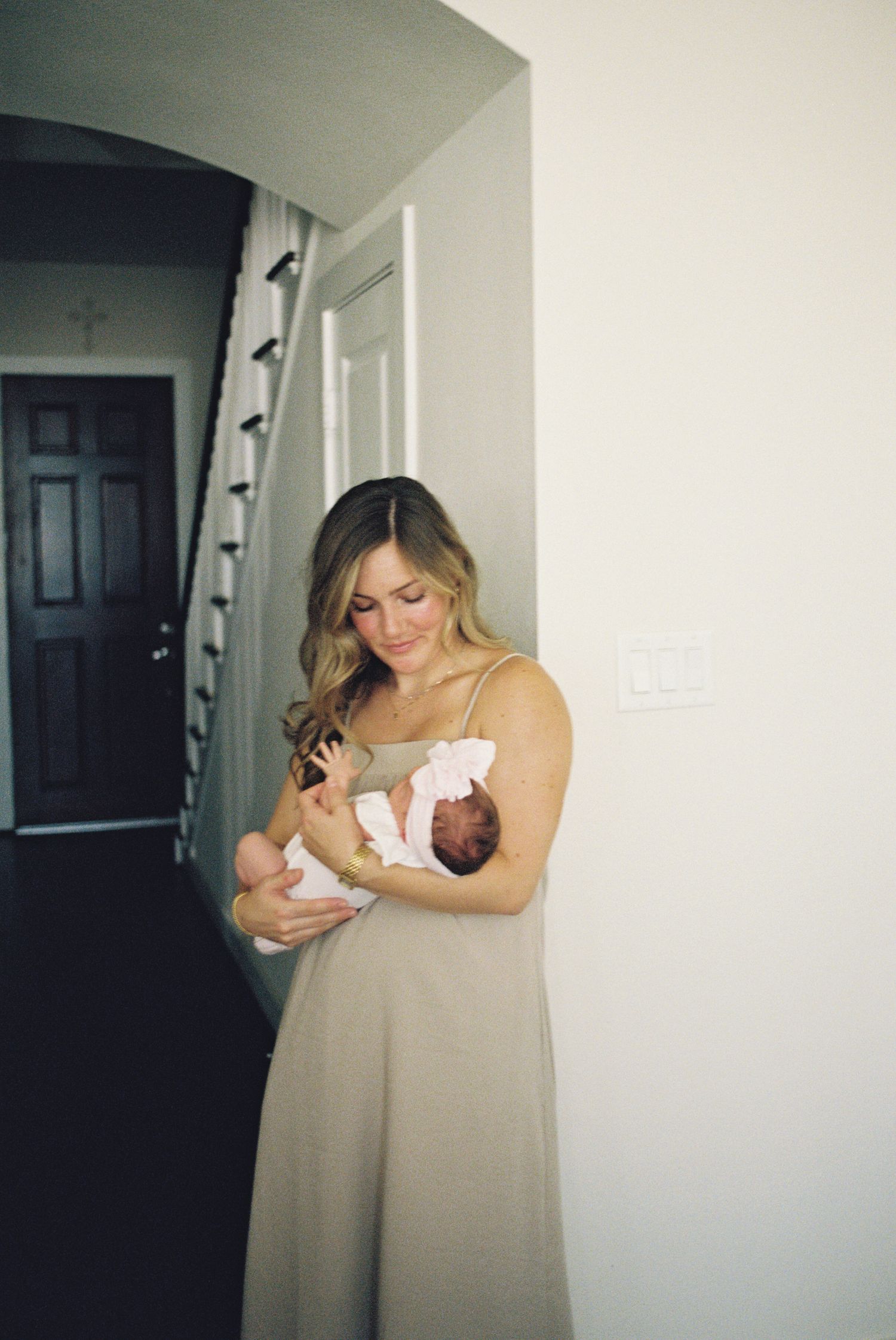 A young mother in a beige dress tenderly holds her newborn baby in a softly lit home entryway.
