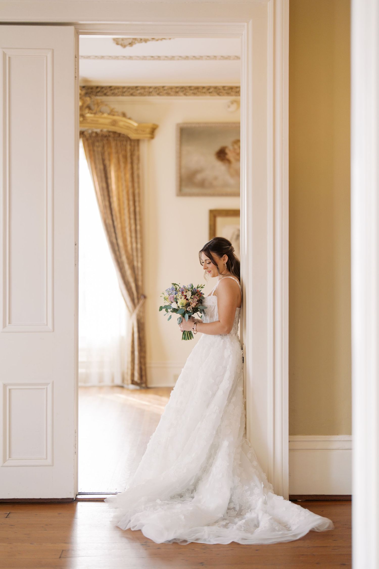A bride in a long, flowing white wedding dress stands in a doorway holding a bouquet of flowers.