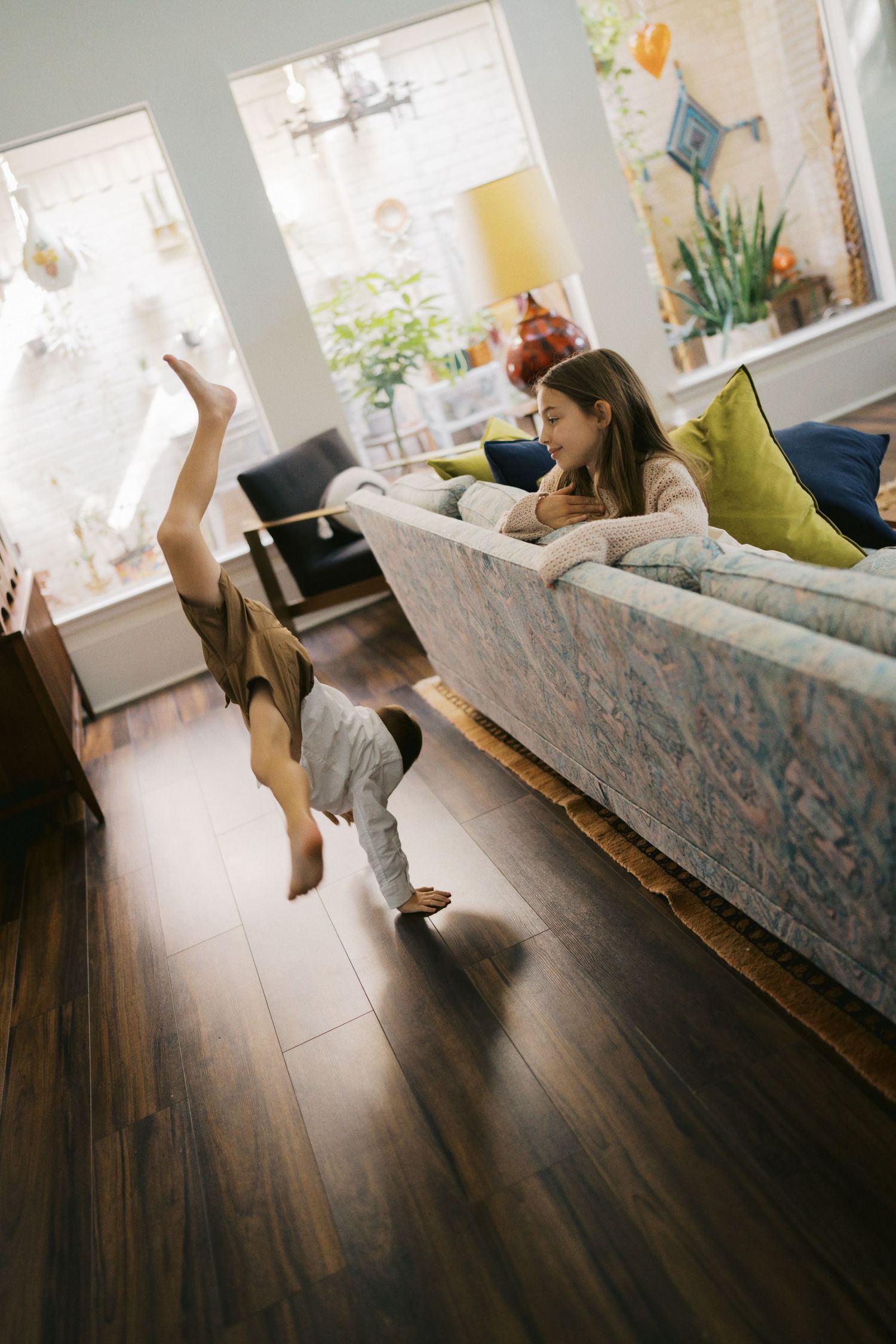 A child does a handstand on hardwood floors while a girl watches from a colorful sofa in a bright living room.