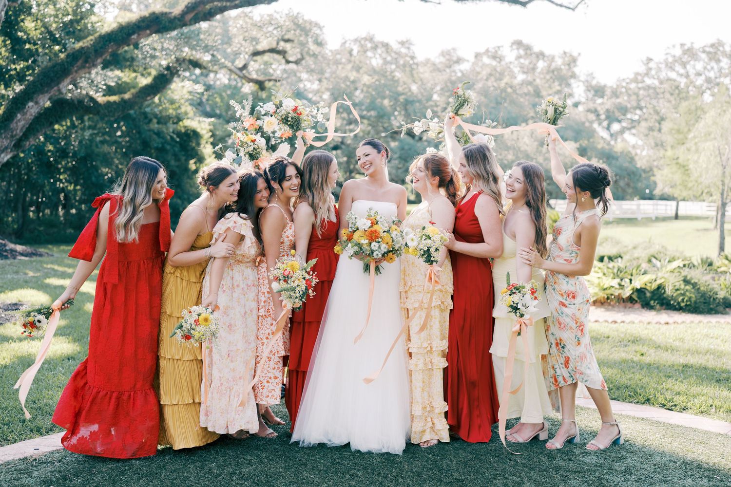 Bridesmaids in red and floral dresses pose joyfully outdoors with the bride.