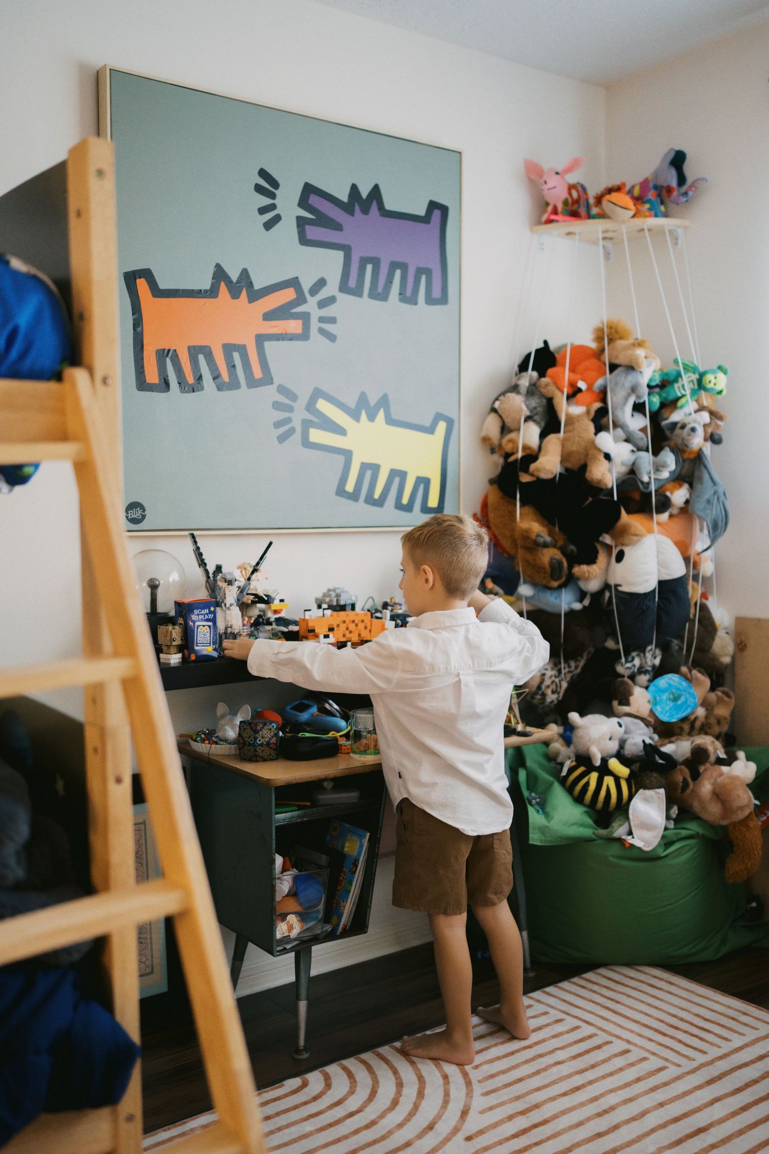 Young boy playing in colorful bedroom with Keith Haring art, toy shelf, and stuffed animal collection.