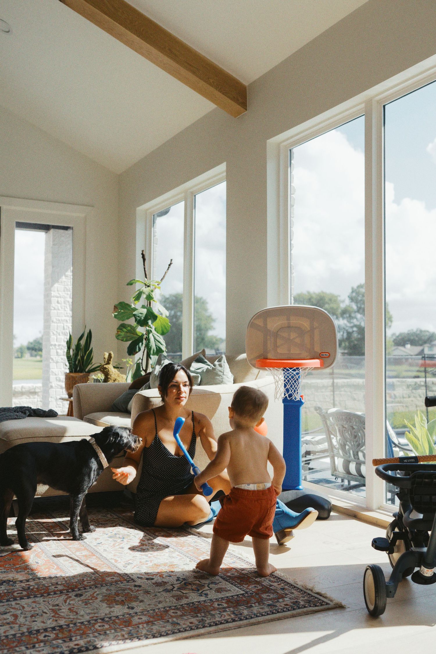Mother playing with toddler and black dog in bright sunlit living room with basketball hoop and large windows.